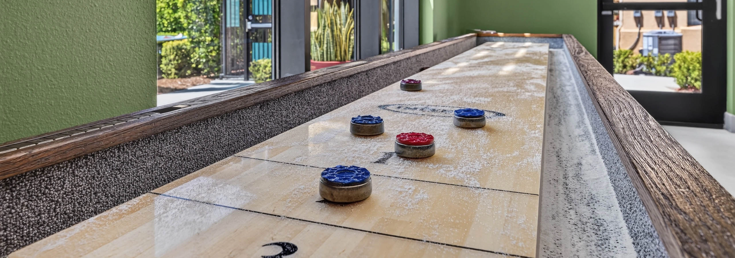 Shuffleboard table at Game Terrace with green walls and large windows overlooking landscaped grounds at AMLI Camarillo apartments.