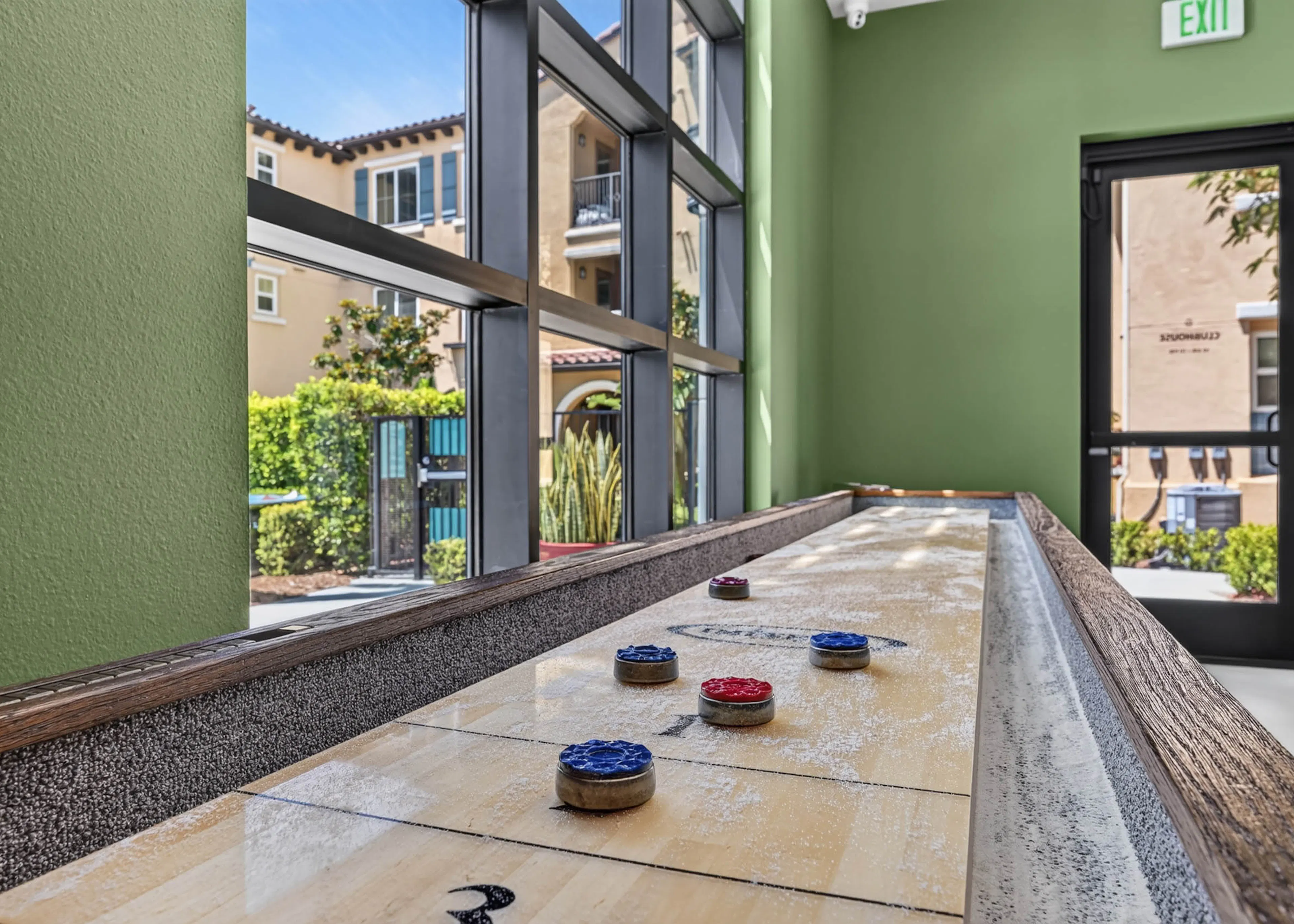 Shuffleboard table at Game Terrace with green walls and large windows overlooking landscaped grounds at AMLI Camarillo apartments.