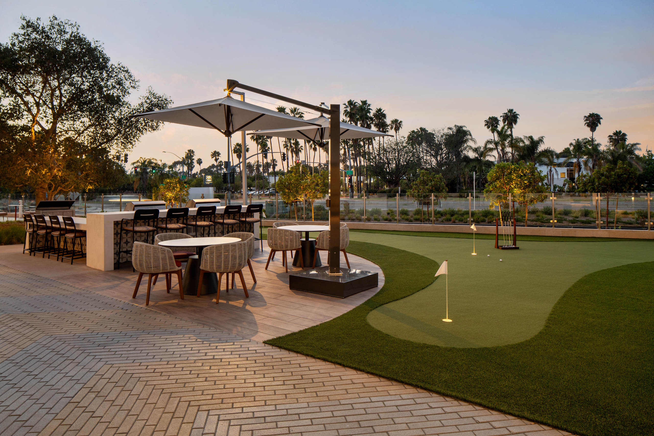 Outdoor gathering space with putting green, bar seating, dining tables, shaded umbrellas, landscaped grounds, palm trees, and evening lighting.
