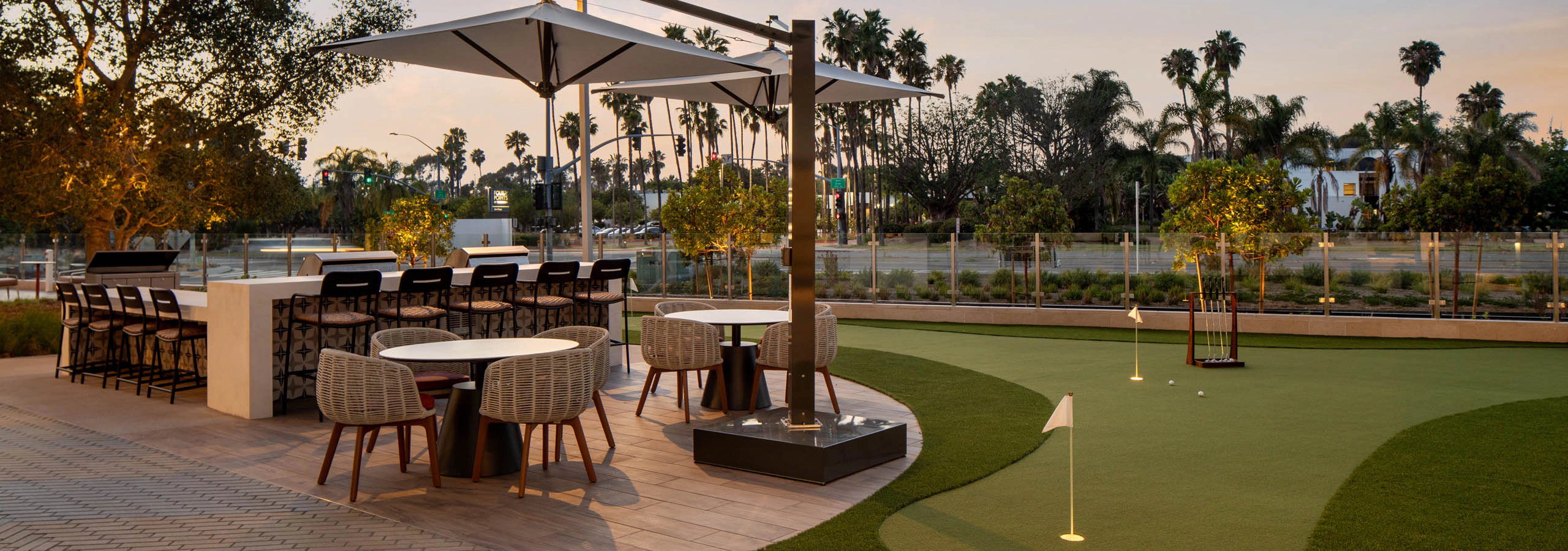 Outdoor gathering space with putting green, bar seating, dining tables, shaded umbrellas, landscaped grounds, palm trees, and evening lighting.