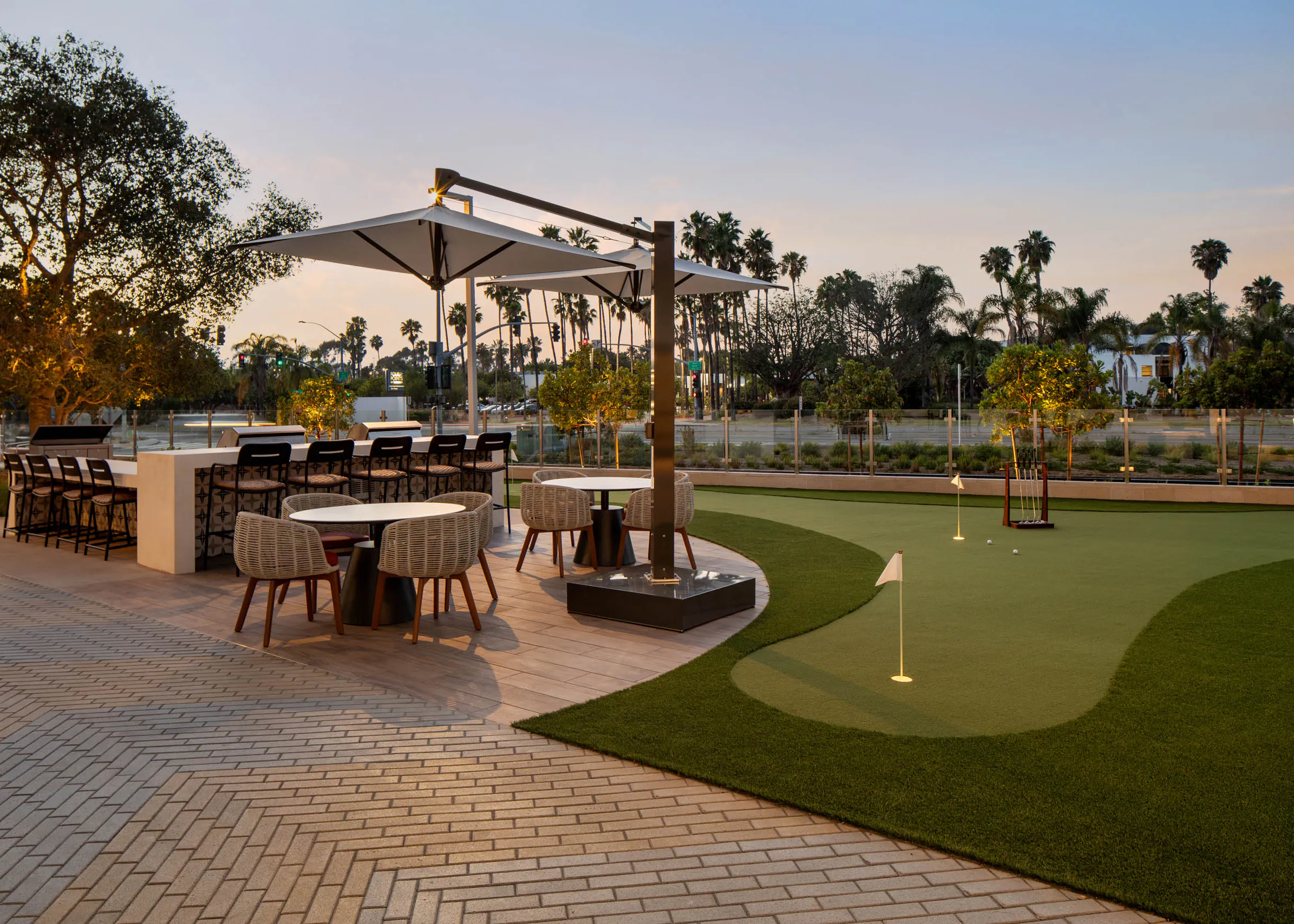 Outdoor gathering space with putting green, bar seating, dining tables, shaded umbrellas, landscaped grounds, palm trees, and evening lighting.