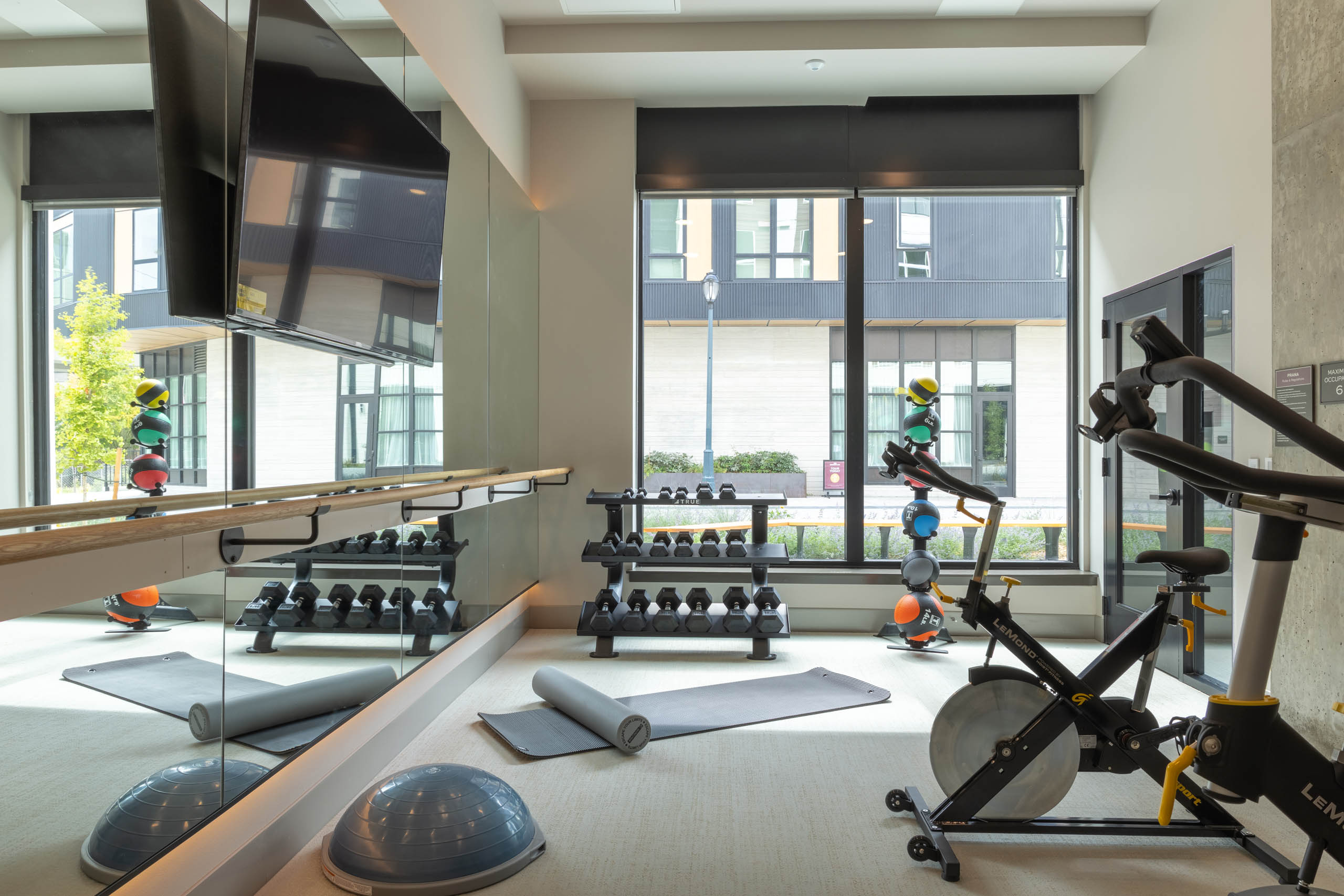 Fitness center studio with a spin bike, dumbbell rack, yoga mats, balance balls, and a barre with mirrored wall.