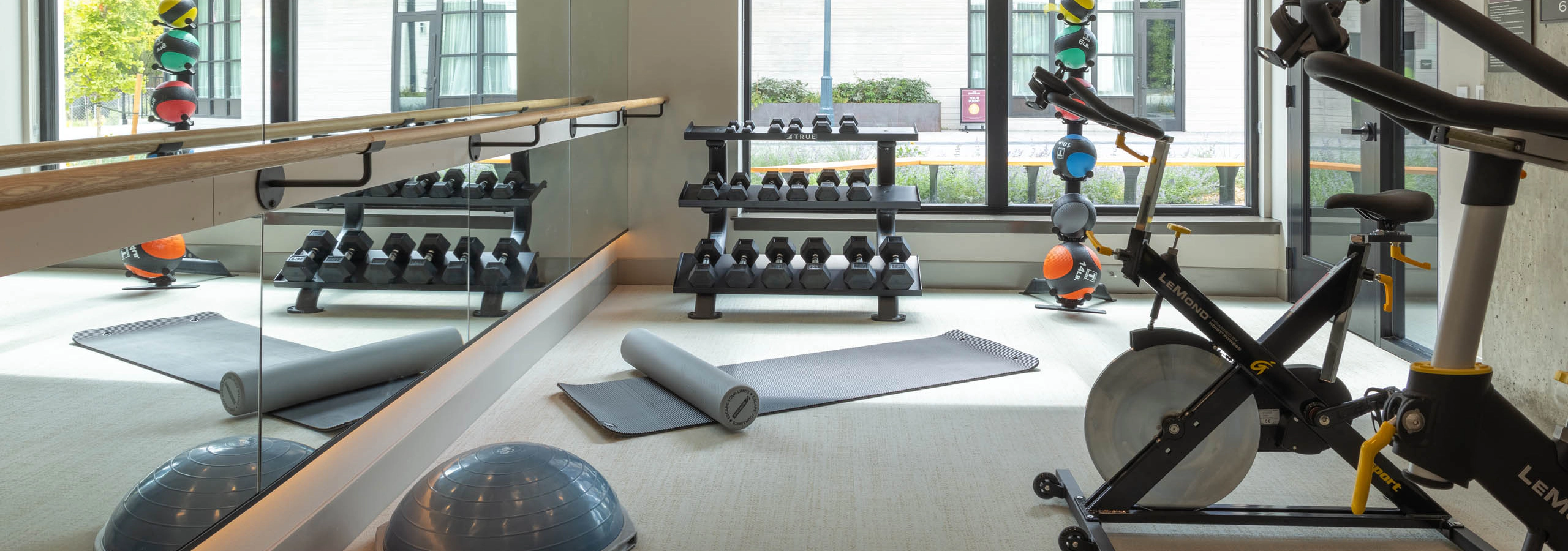 Fitness center studio with a spin bike, dumbbell rack, yoga mats, balance balls, and a barre with mirrored wall.