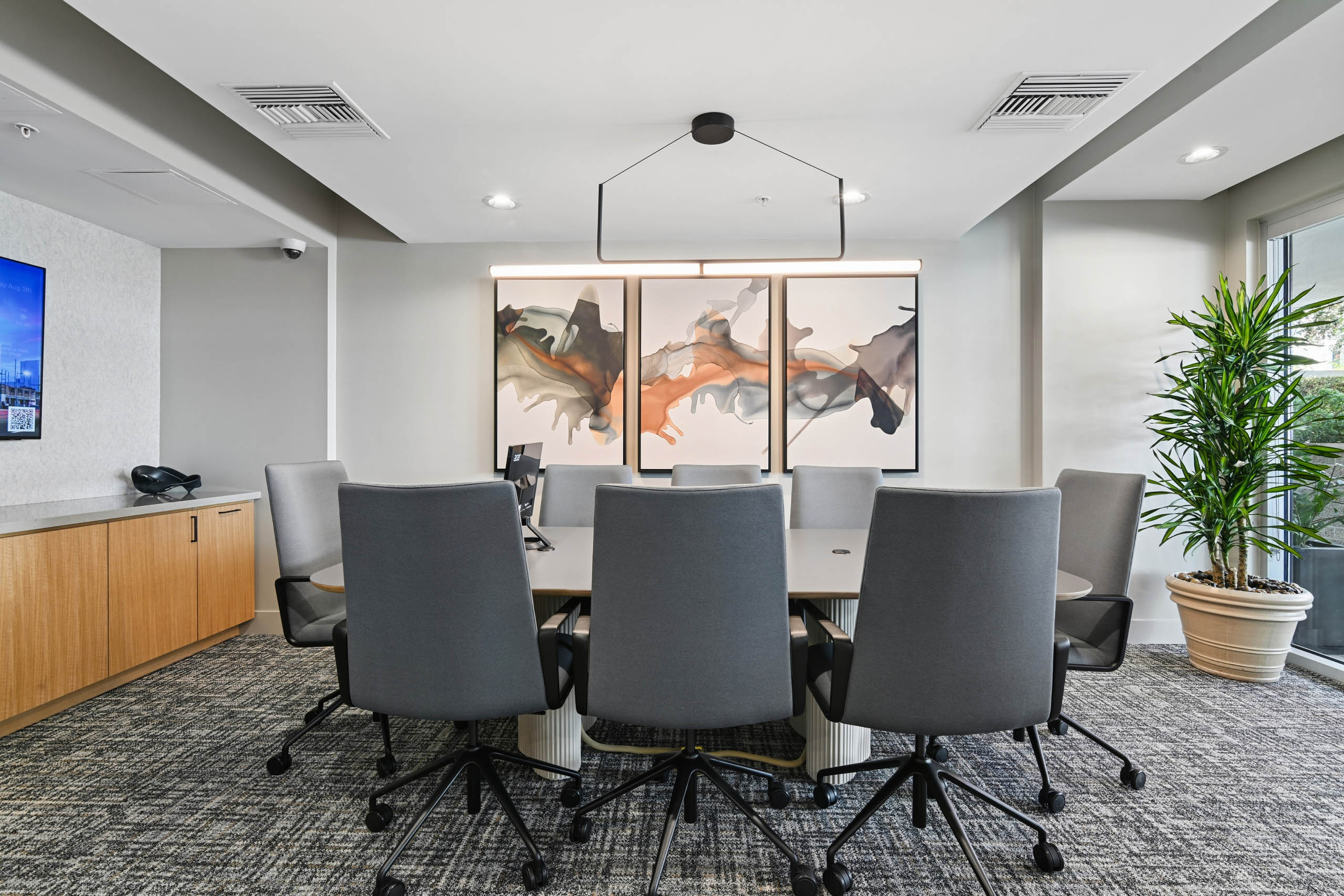 Conference room at AMLI Lex on Orange with a large table, gray rolling chairs, a modern linear pendant light, abstract wall art, a wall-mounted screen, wood cabinetry, and a tall potted plant by the window.