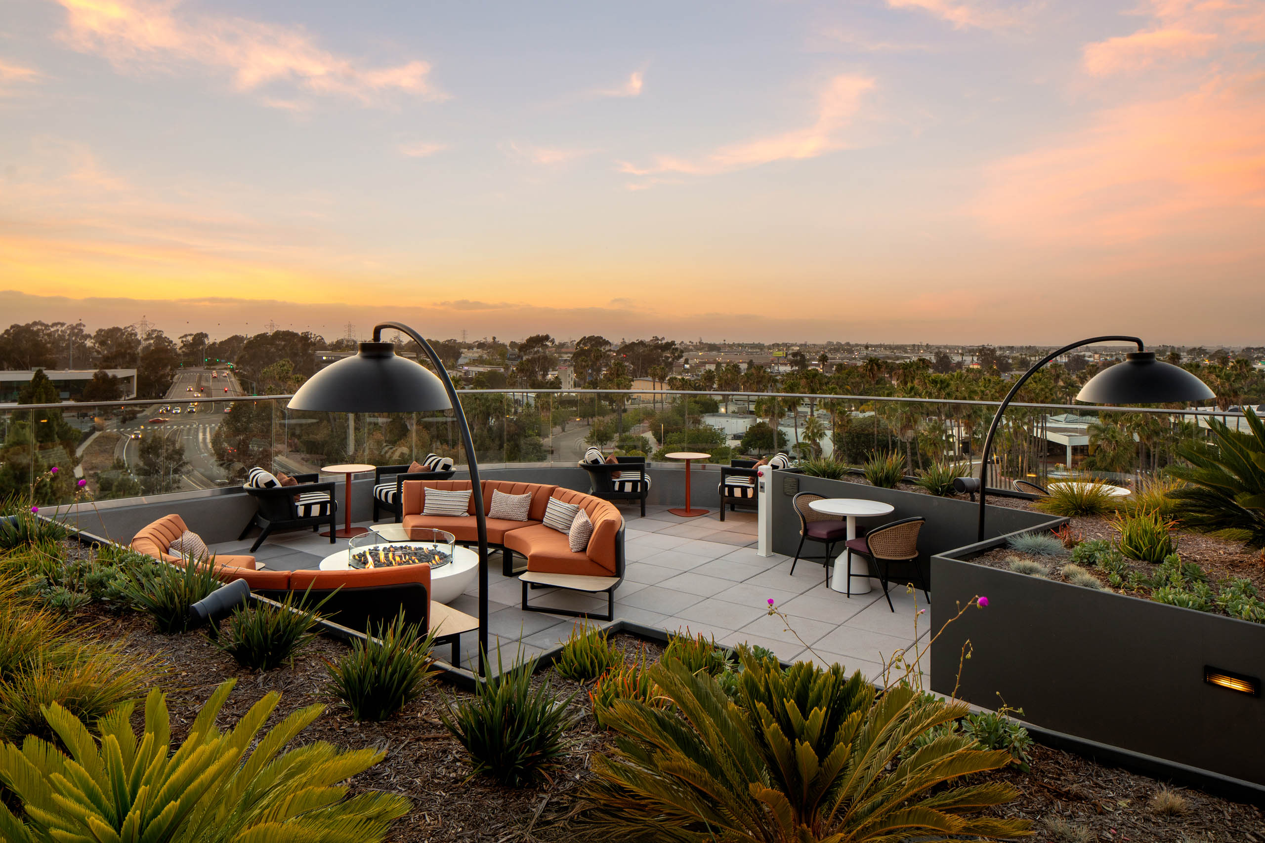 Rooftop lounge at sunset with outdoor seating, fire pit tables, landscaped planters, overhead lighting, and panoramic views beyond glass railings.