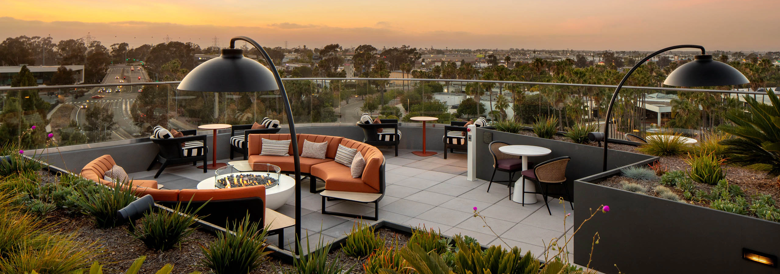 Rooftop lounge at sunset with outdoor seating, fire pit tables, landscaped planters, overhead lighting, and panoramic views beyond glass railings.