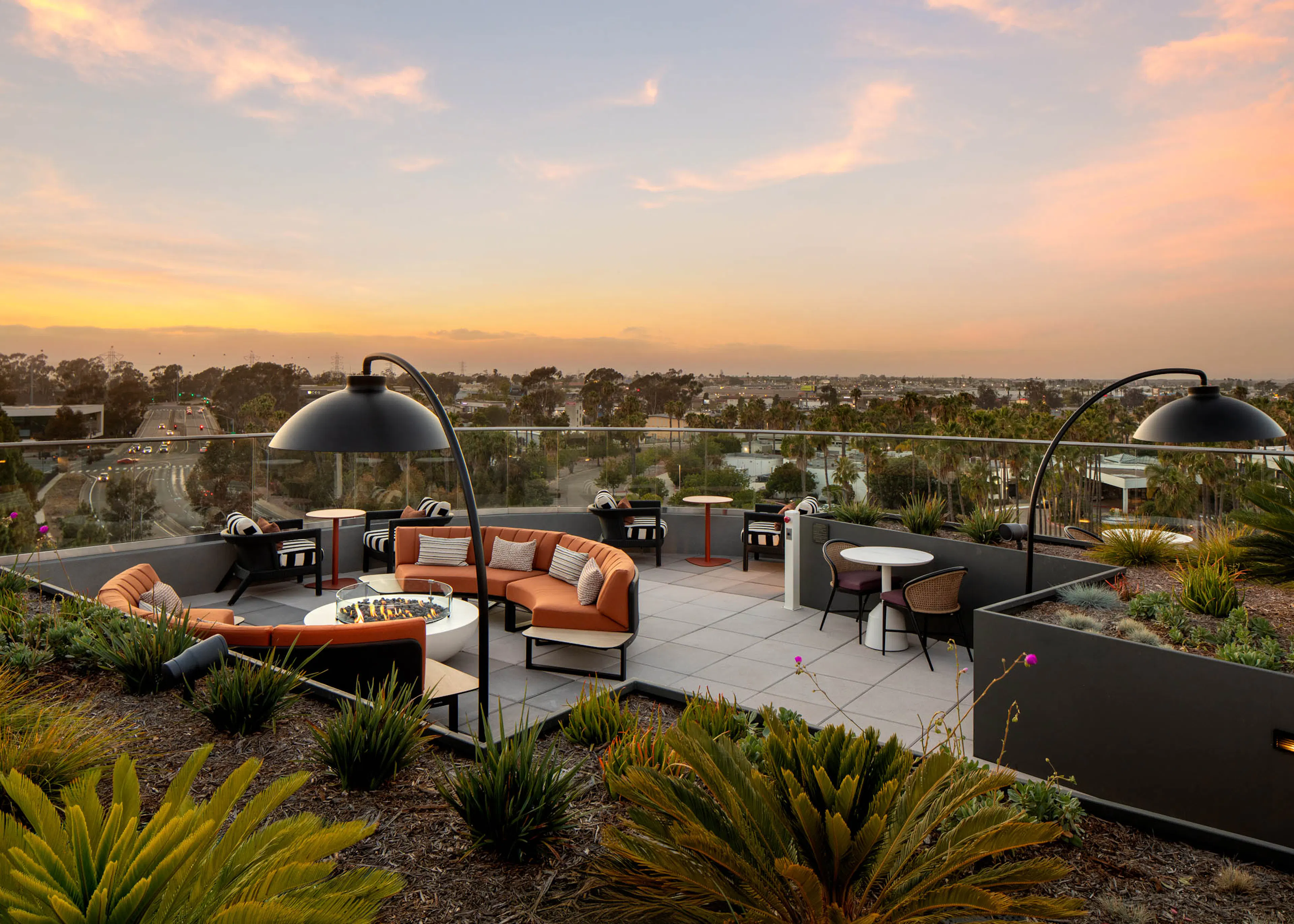 Rooftop lounge at sunset with outdoor seating, fire pit tables, landscaped planters, overhead lighting, and panoramic views beyond glass railings.