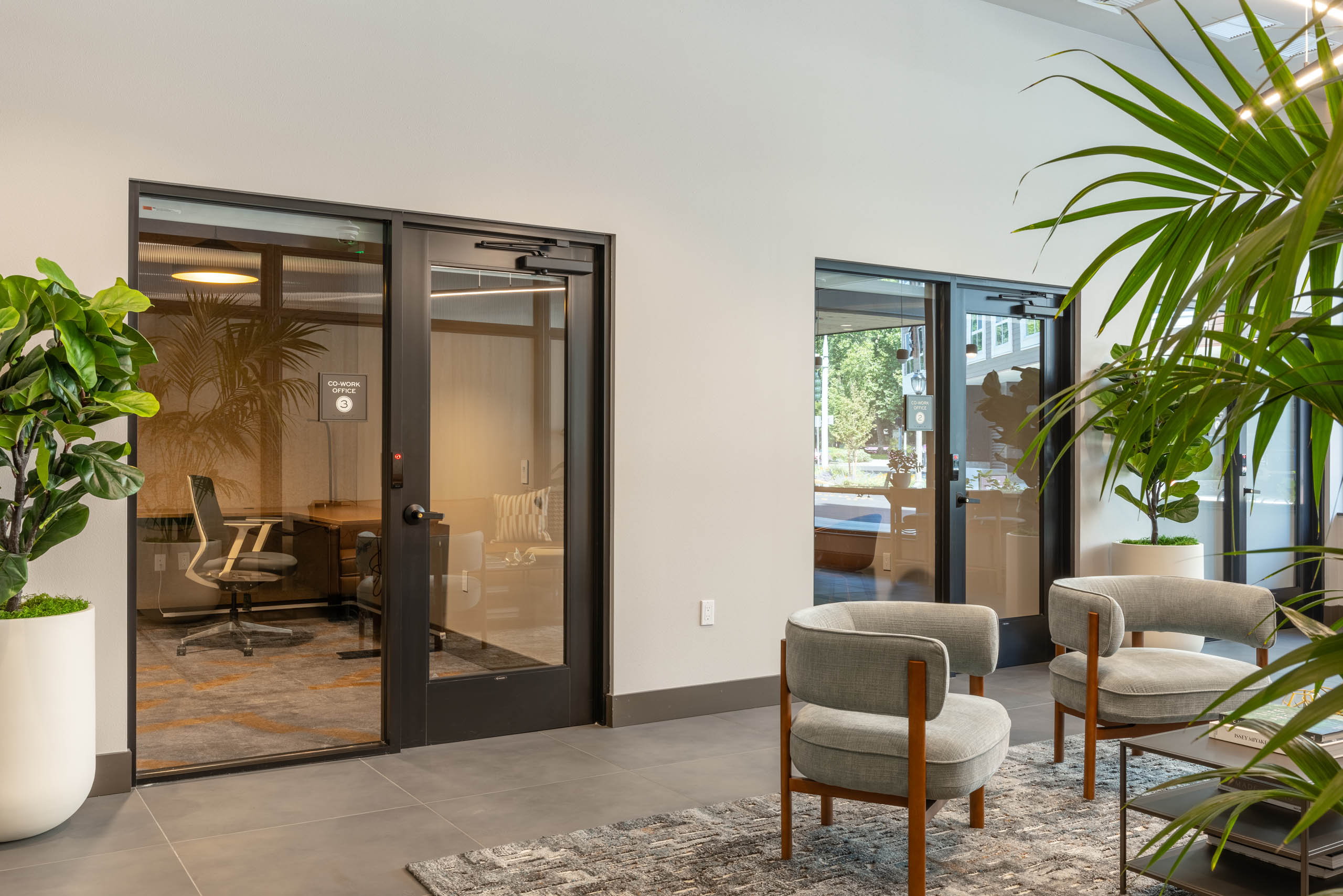 East building co-work office suites with glass-front private offices, desk and lounge seating visible inside, plus a shared lounge area with armchairs and greenery in the hallway.