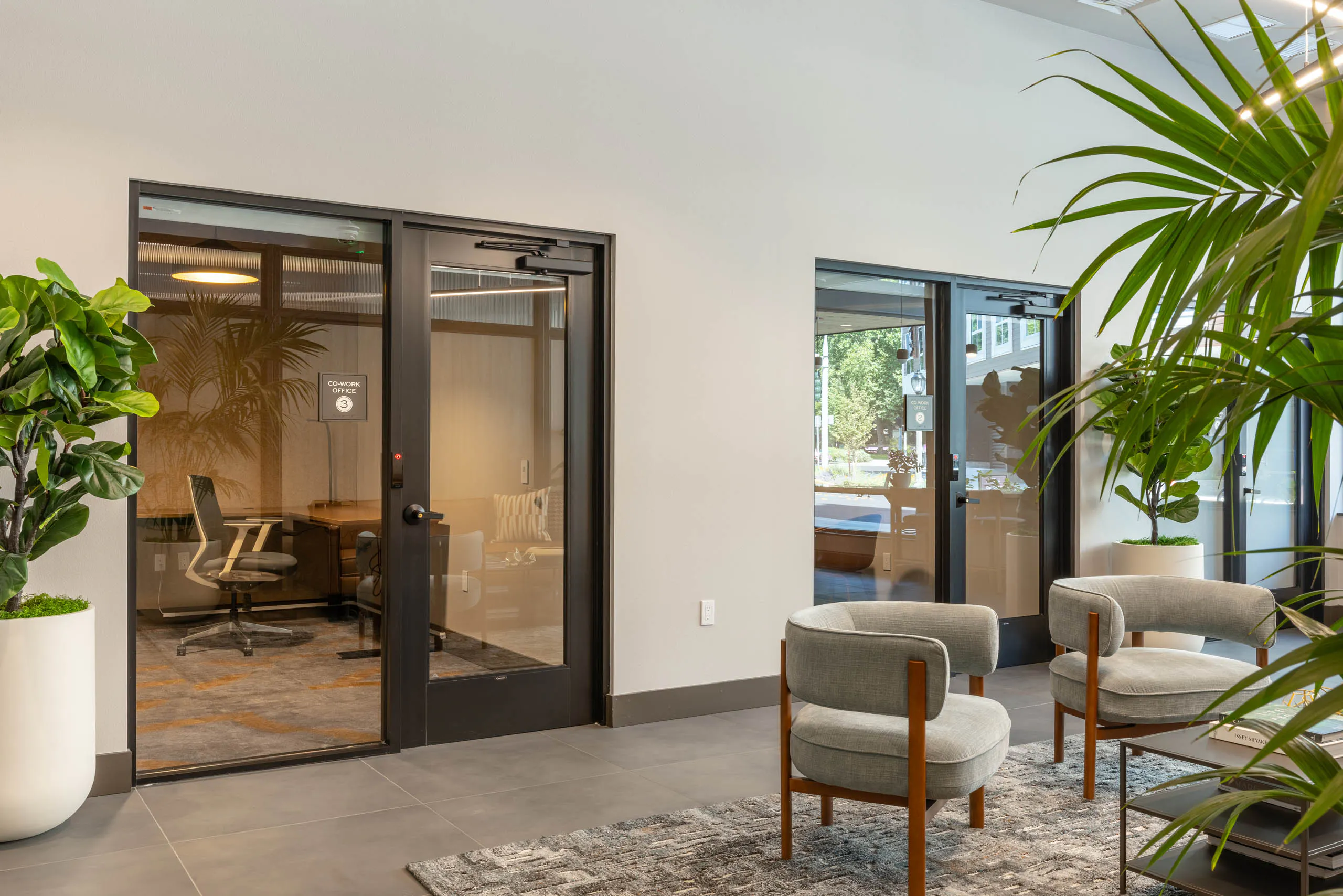 East building co-work office suites with glass-front private offices, desk and lounge seating visible inside, plus a shared lounge area with armchairs and greenery in the hallway.