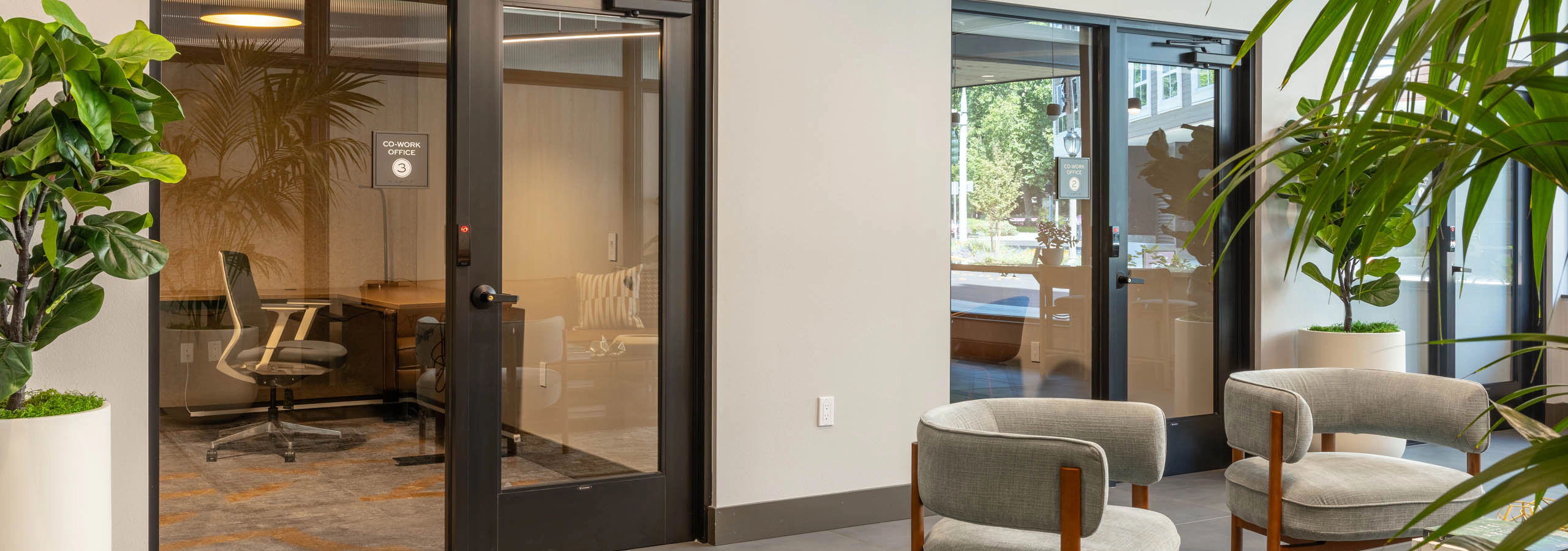 East building co-work office suites with glass-front private offices, desk and lounge seating visible inside, plus a shared lounge area with armchairs and greenery in the hallway.