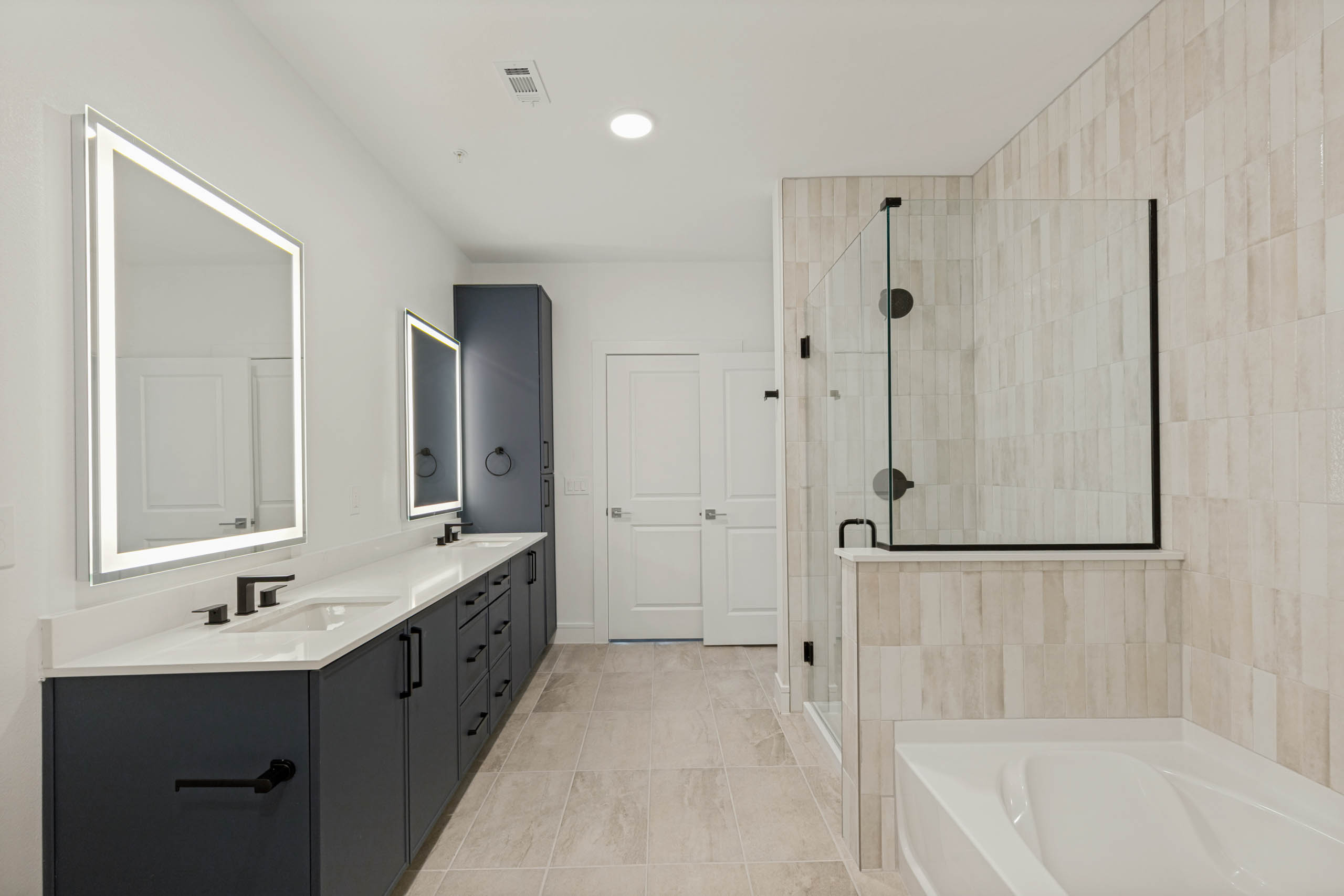 Beautifual bathroom with double vanity, illuminated mirrors, quartz countertops, glass-enclosed shower, soaking tub, and tile flooring.