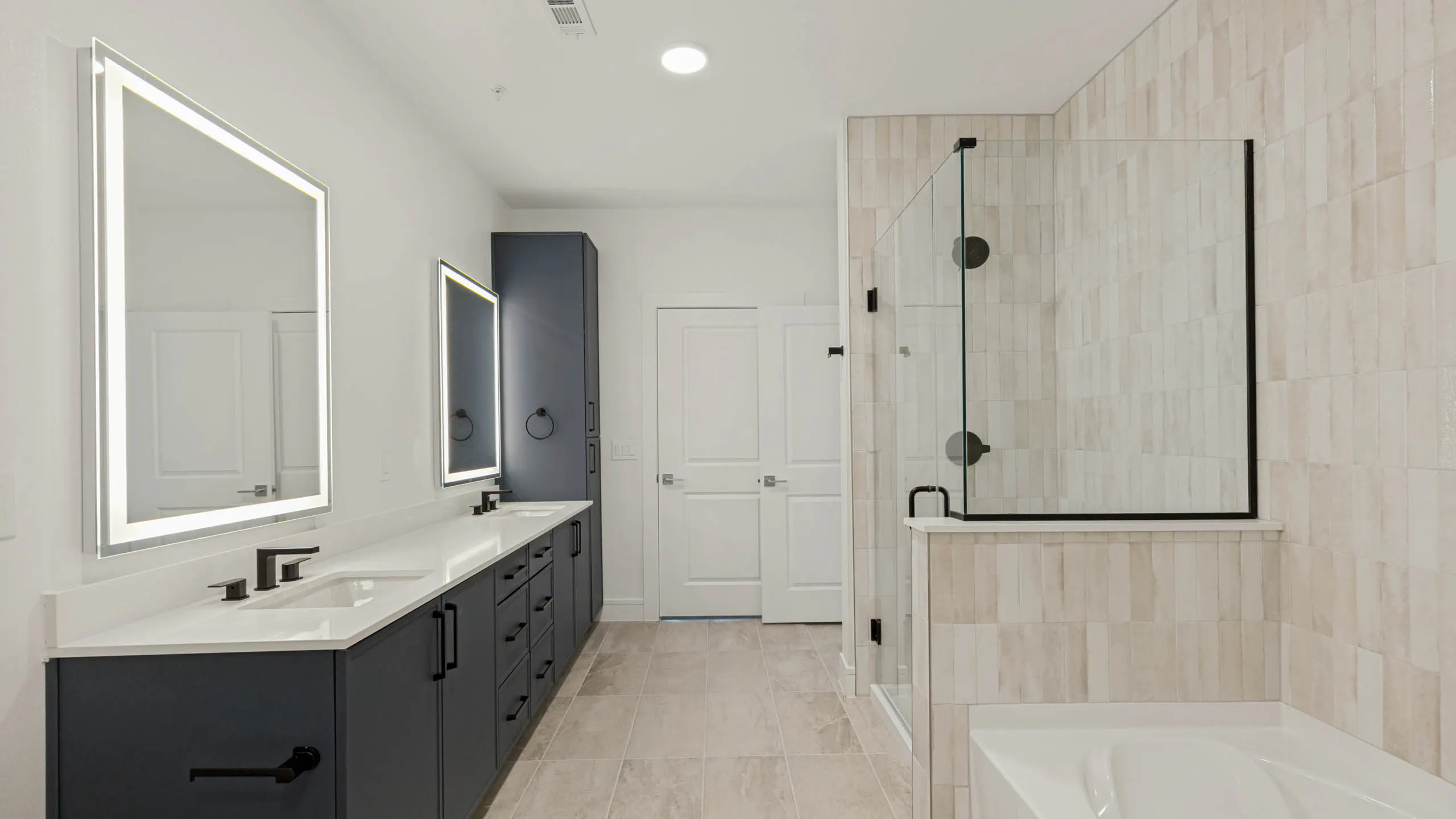 Modern bathroom with double vanity, illuminated mirrors, quartz countertops, glass-enclosed shower, soaking tub, and tile flooring.