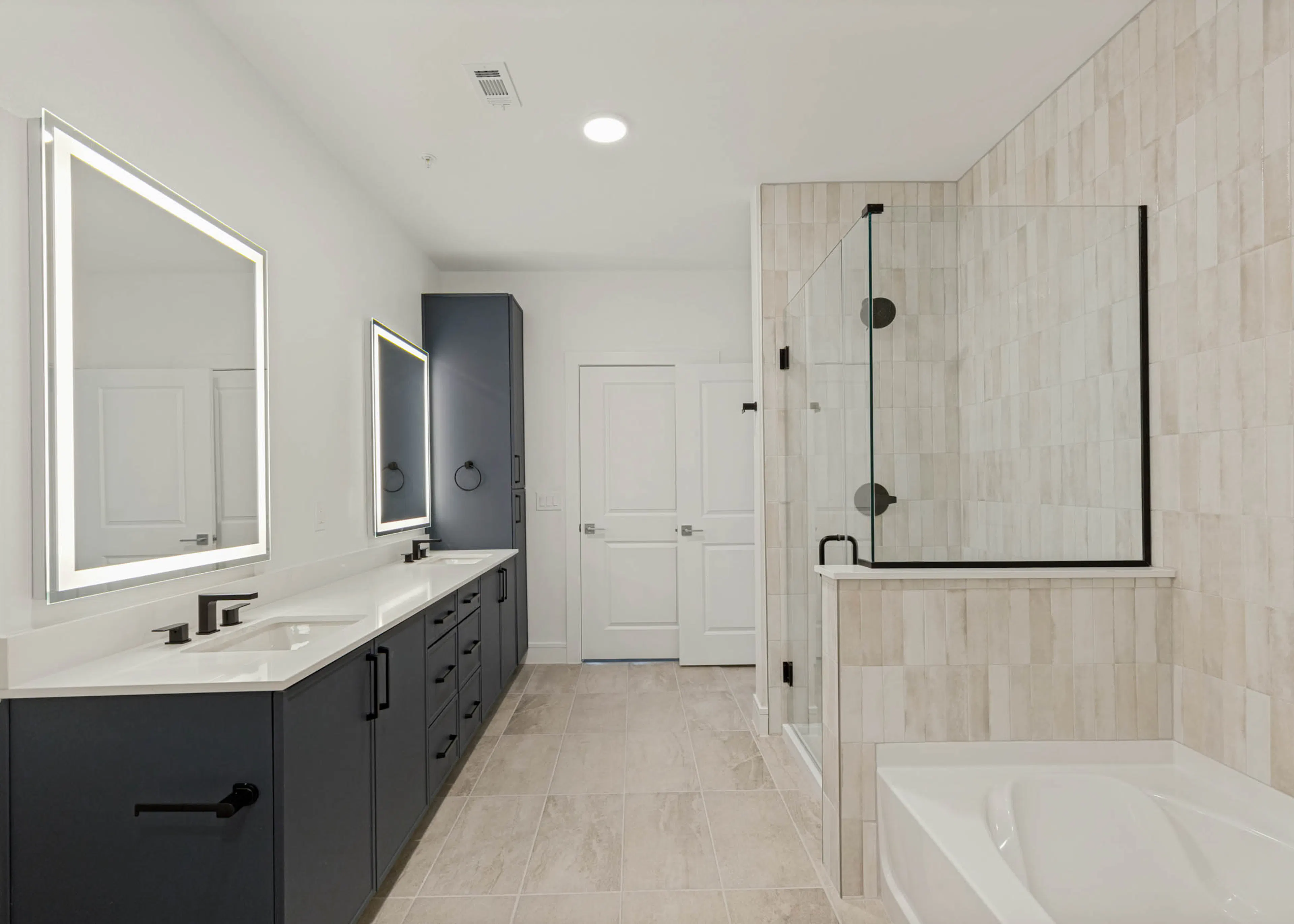 Modern bathroom with double vanity, illuminated mirrors, quartz countertops, glass-enclosed shower, soaking tub, and tile flooring.