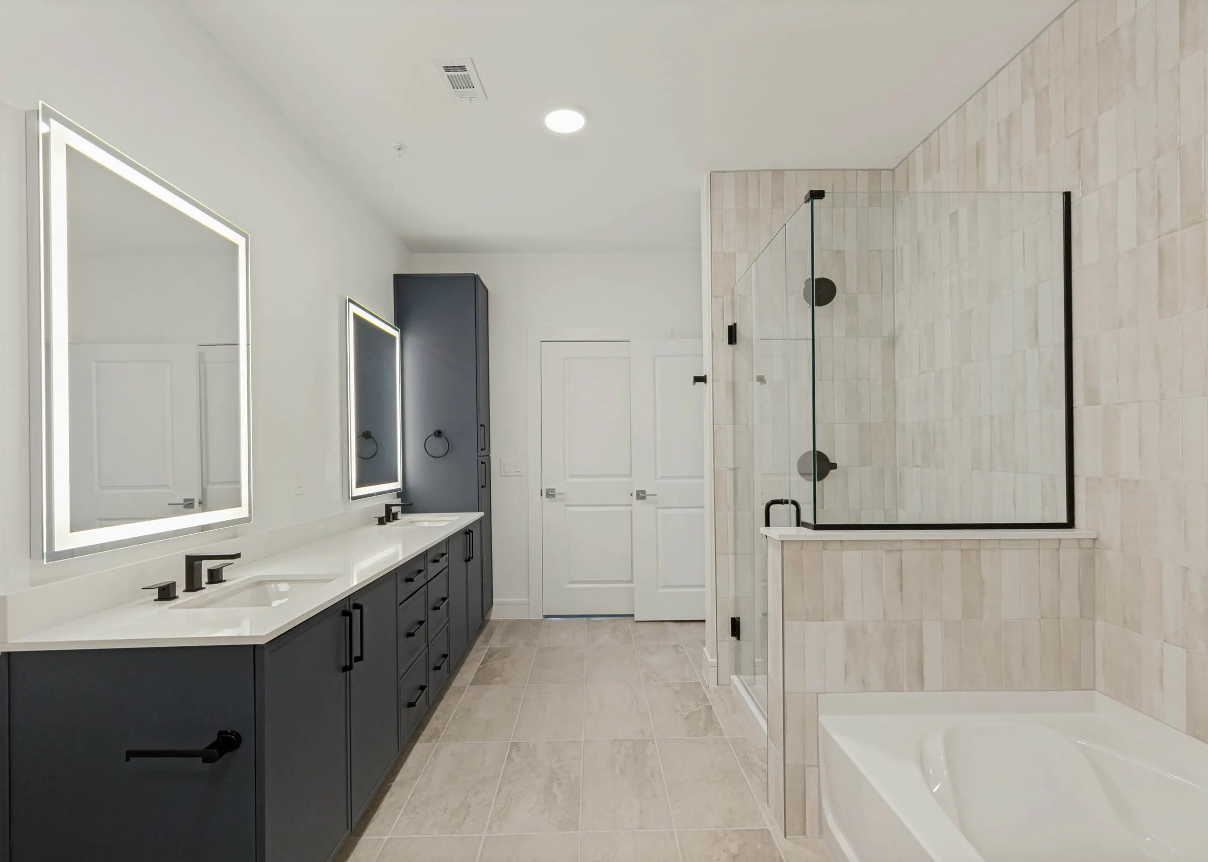 Modern bathroom with double vanity, illuminated mirrors, quartz countertops, glass-enclosed shower, soaking tub, and tile flooring.