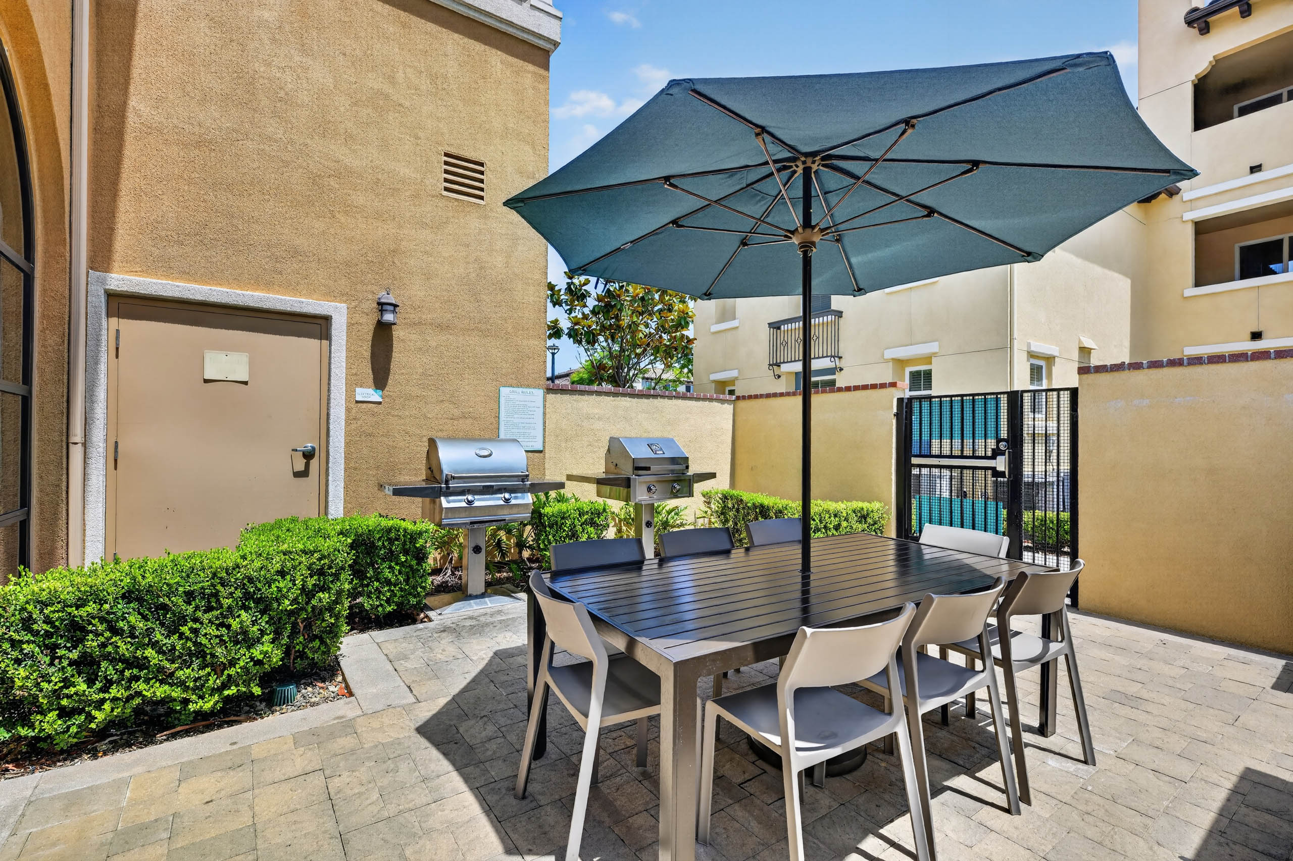 Outdoor grilling area at AMLI Camarillo apartments with barbecue grills, dining table, chairs, large blue umbrella and green hedges.