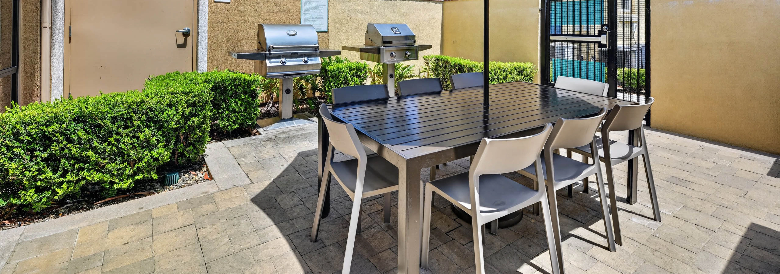 Outdoor grilling area at AMLI Camarillo apartments with barbecue grills, dining table, chairs, large blue umbrella and green hedges.
