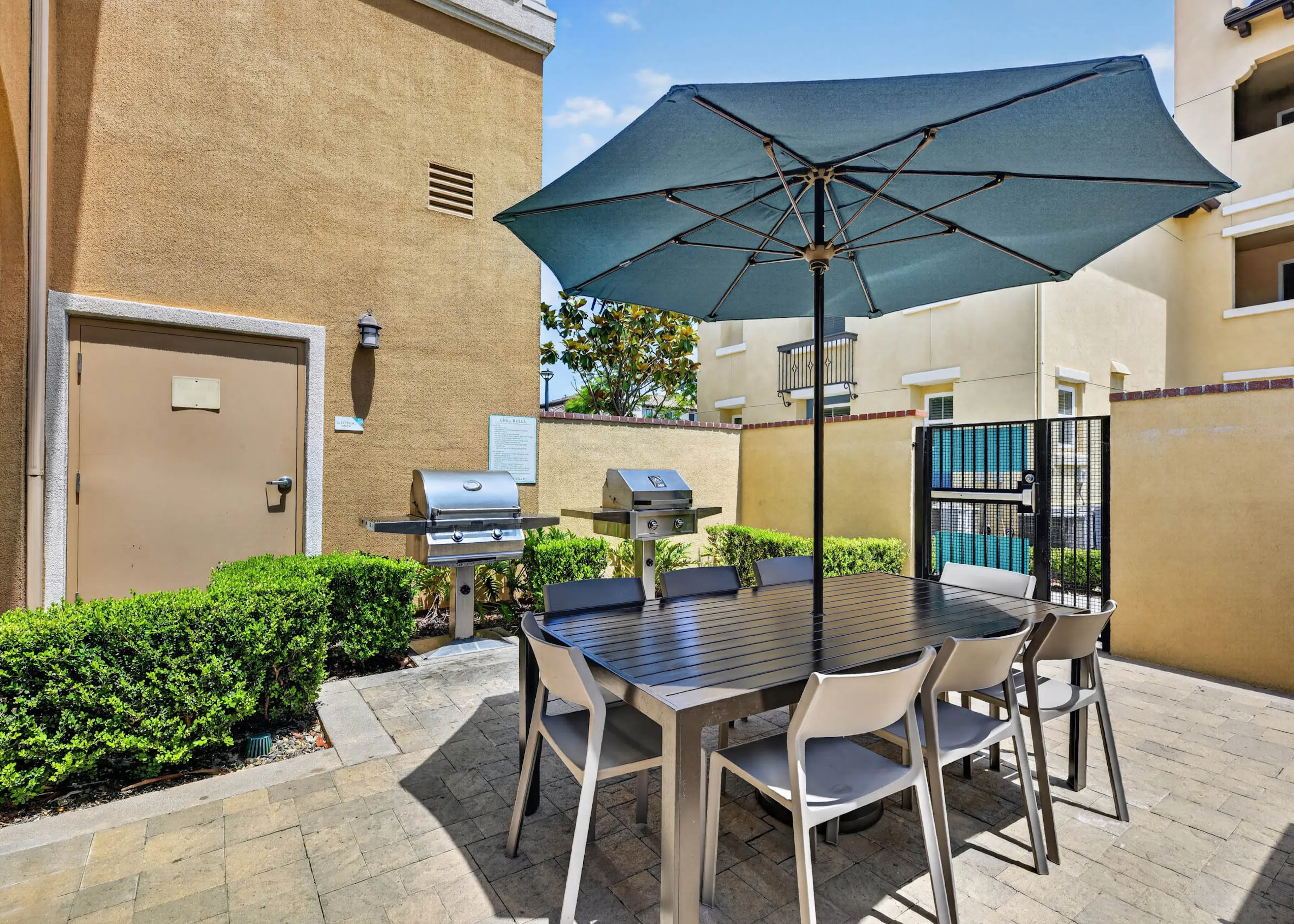 Outdoor grilling area at AMLI Camarillo apartments with barbecue grills, dining table, chairs, large blue umbrella and green hedges.