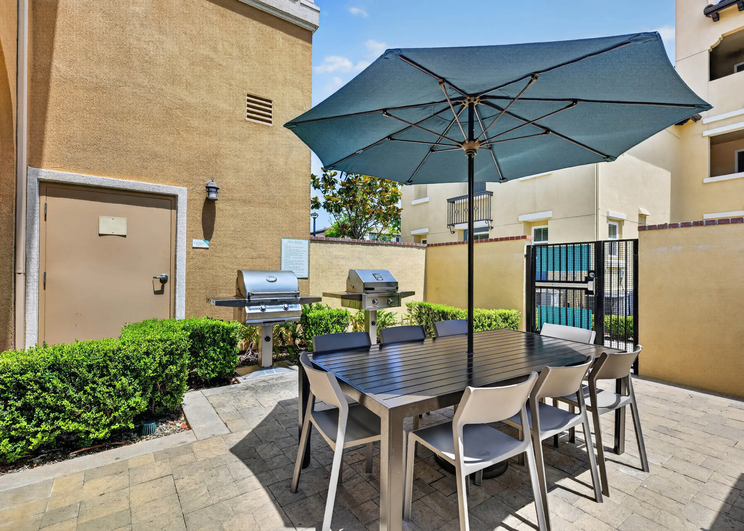 Outdoor grilling area at AMLI Camarillo apartments with barbecue grills, dining table, chairs, large blue umbrella and green hedges.