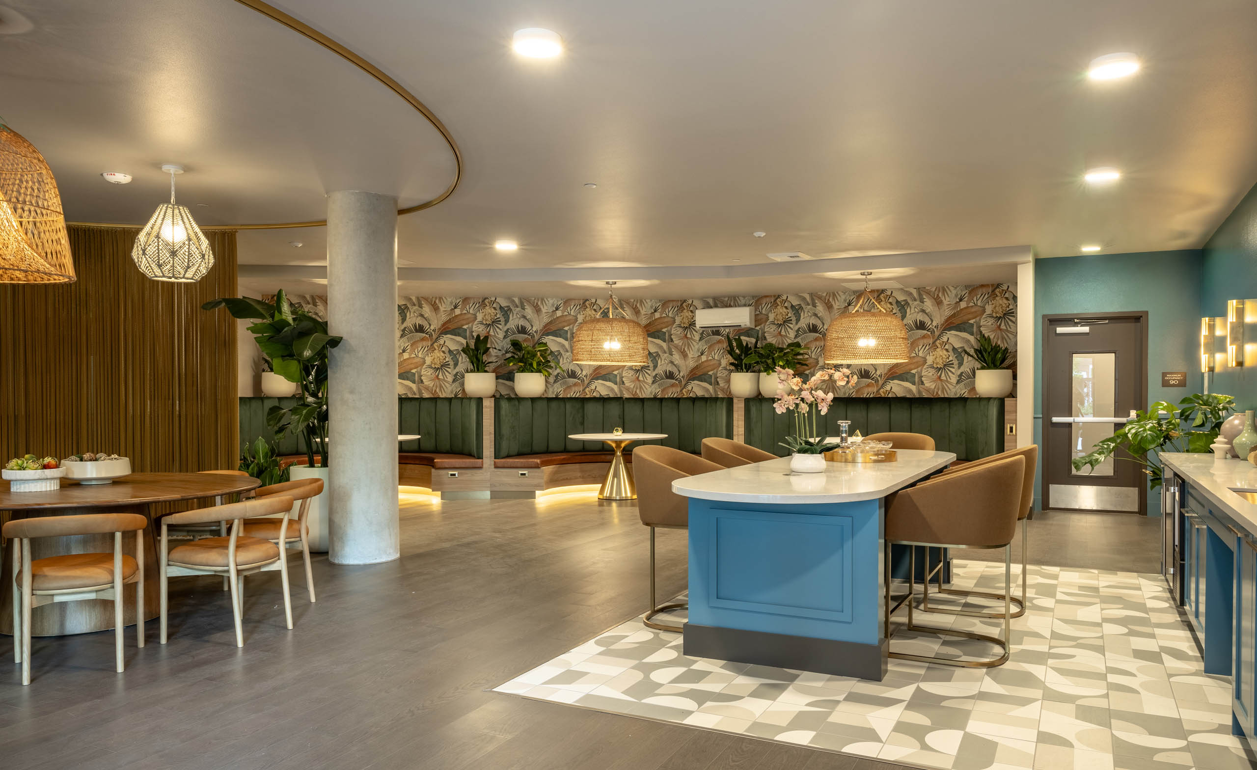Havana Lounge community room featuring a blue kitchen island with bar seating, tropical wallpaper, woven pendant lights, and lounge seating throughout.