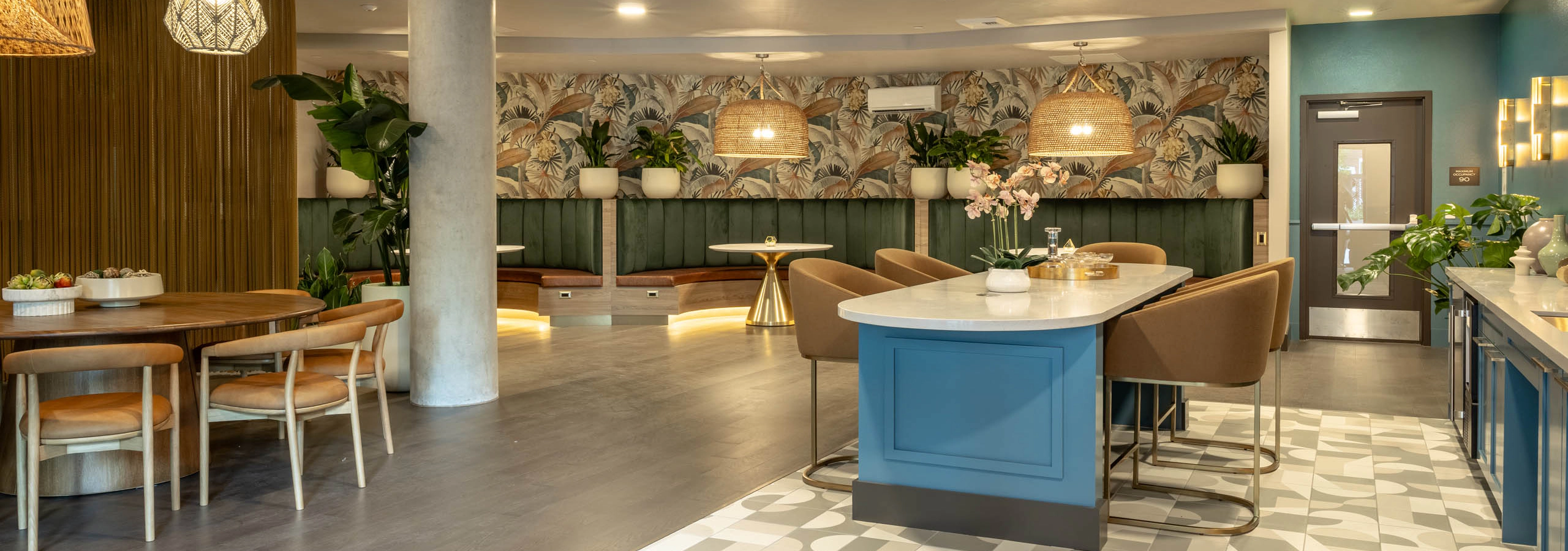 Havana Lounge community room featuring a blue kitchen island with bar seating, tropical wallpaper, woven pendant lights, and lounge seating throughout.
