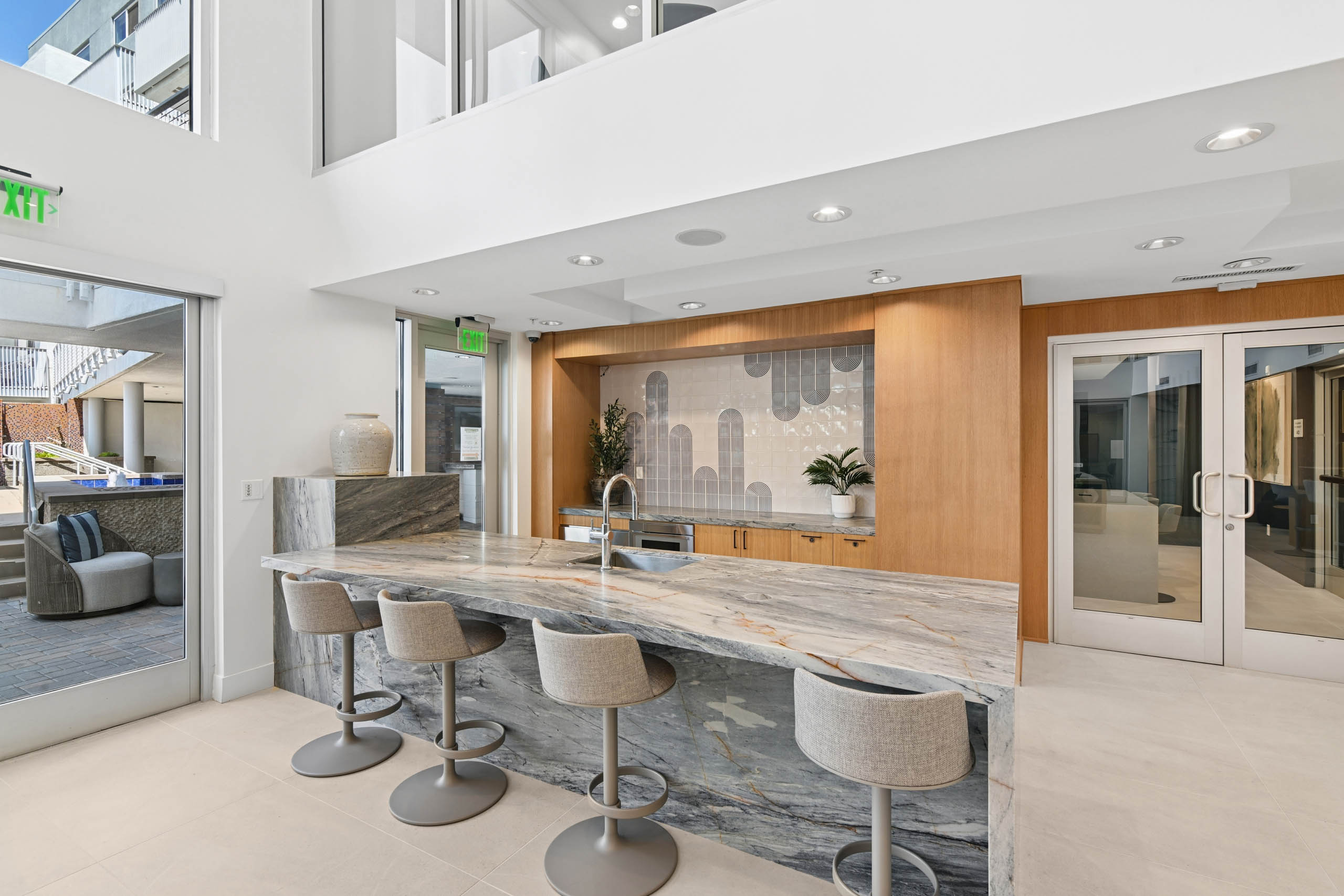 Clubroom kitchen at AMLI Lex on Orange with a stone counter with sink and upholstered bar stools, wood cabinetry, a patterned tile backsplash, and glass doors opening to an outdoor seating area.