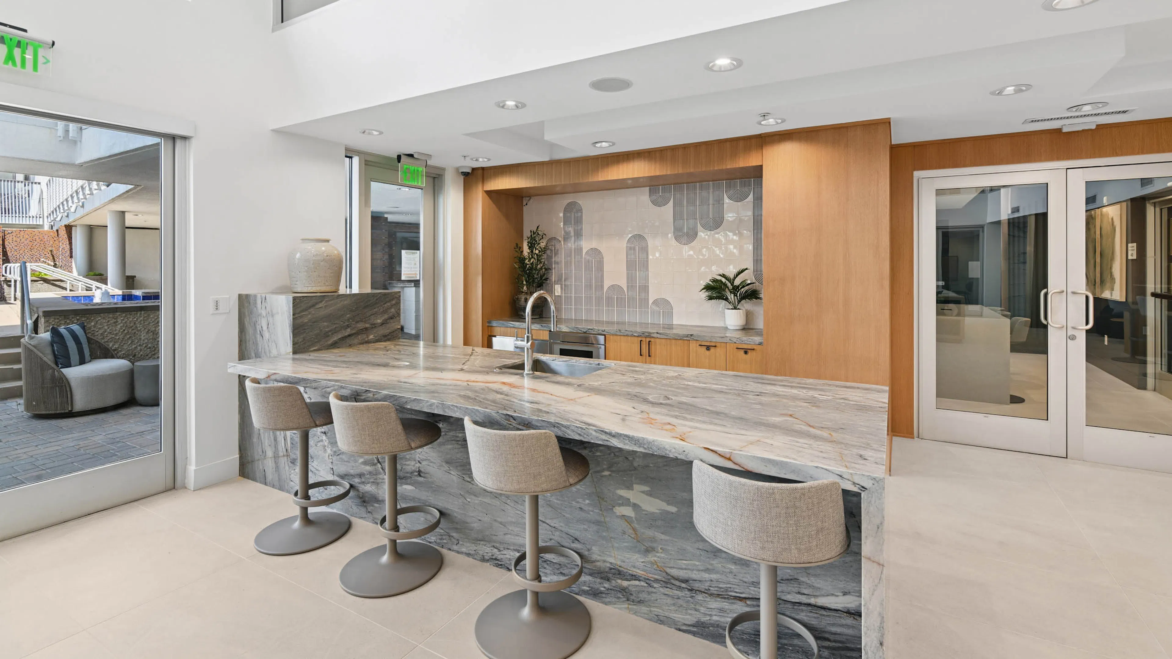 Clubroom kitchen at AMLI Lex on Orange with a stone counter with sink and upholstered bar stools, wood cabinetry, a patterned tile backsplash, and glass doors opening to an outdoor seating area.