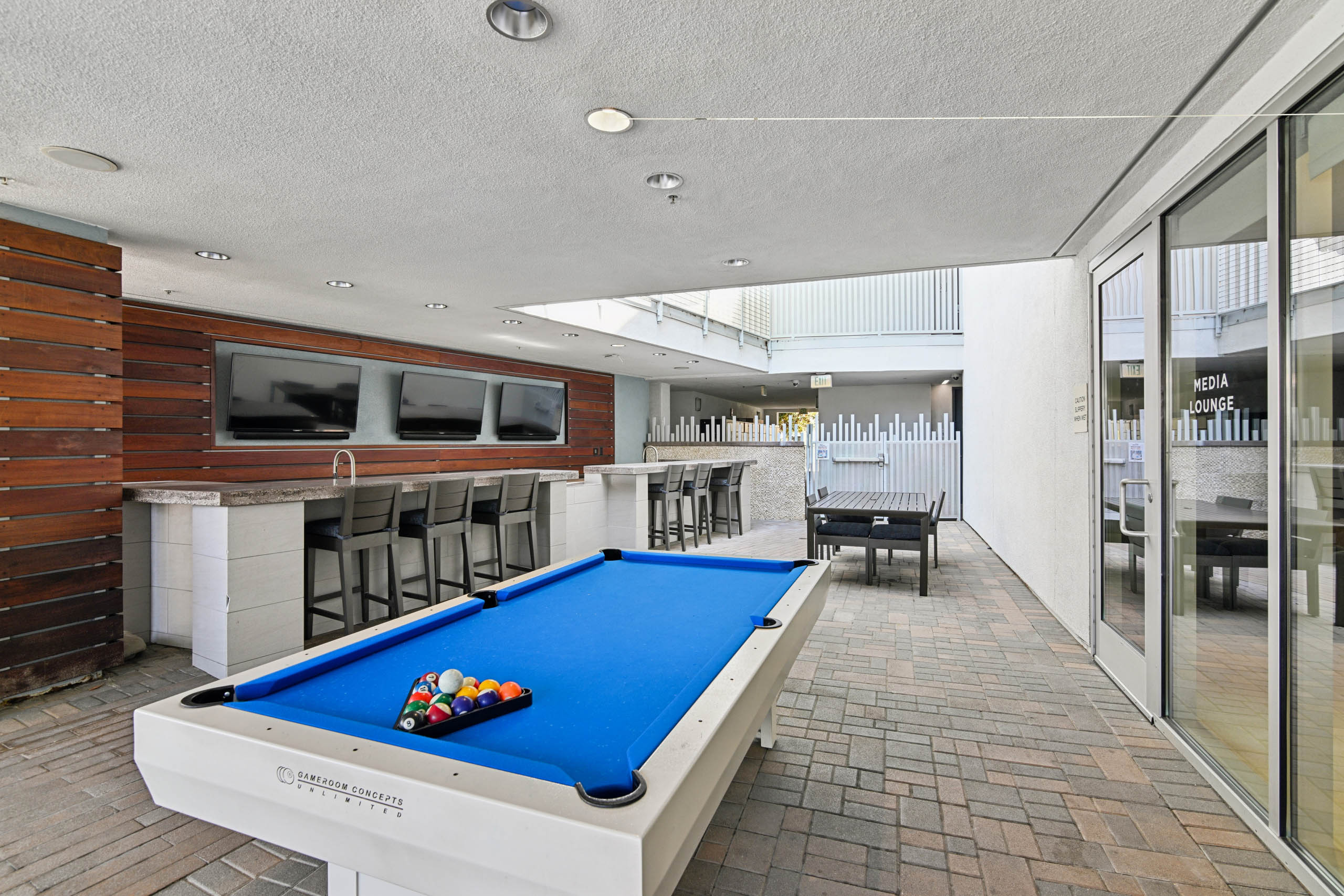 Covered outdoor billiards and bar area at AMLI Lex on Orange with a blue pool table, a bar counter with stools, three wall-mounted TVs on wood paneling, and an outdoor dining table.