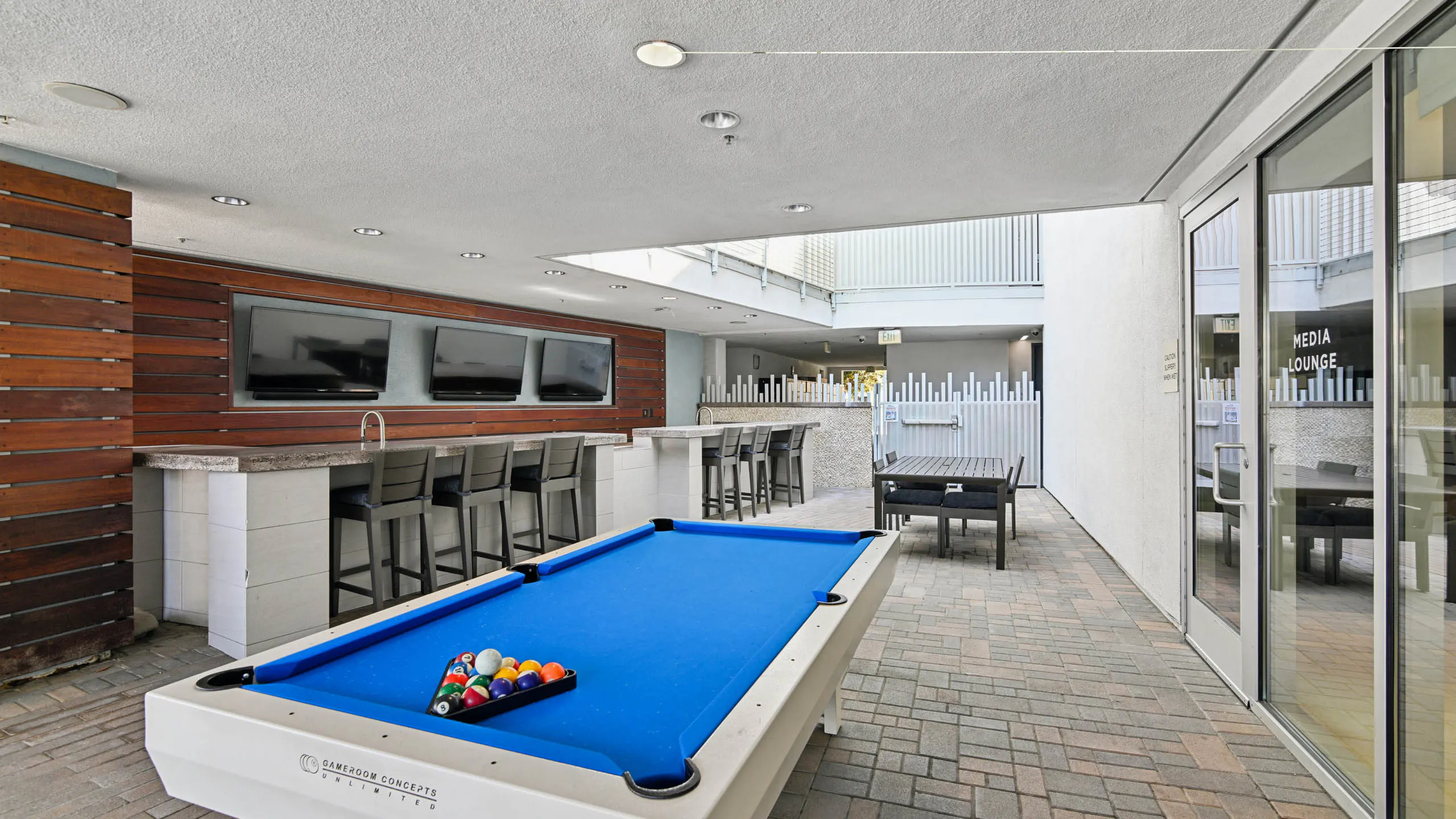 Covered outdoor billiards and bar area at AMLI Lex on Orange with a blue pool table, a bar counter with stools, three wall-mounted TVs on wood paneling, and an outdoor dining table.
