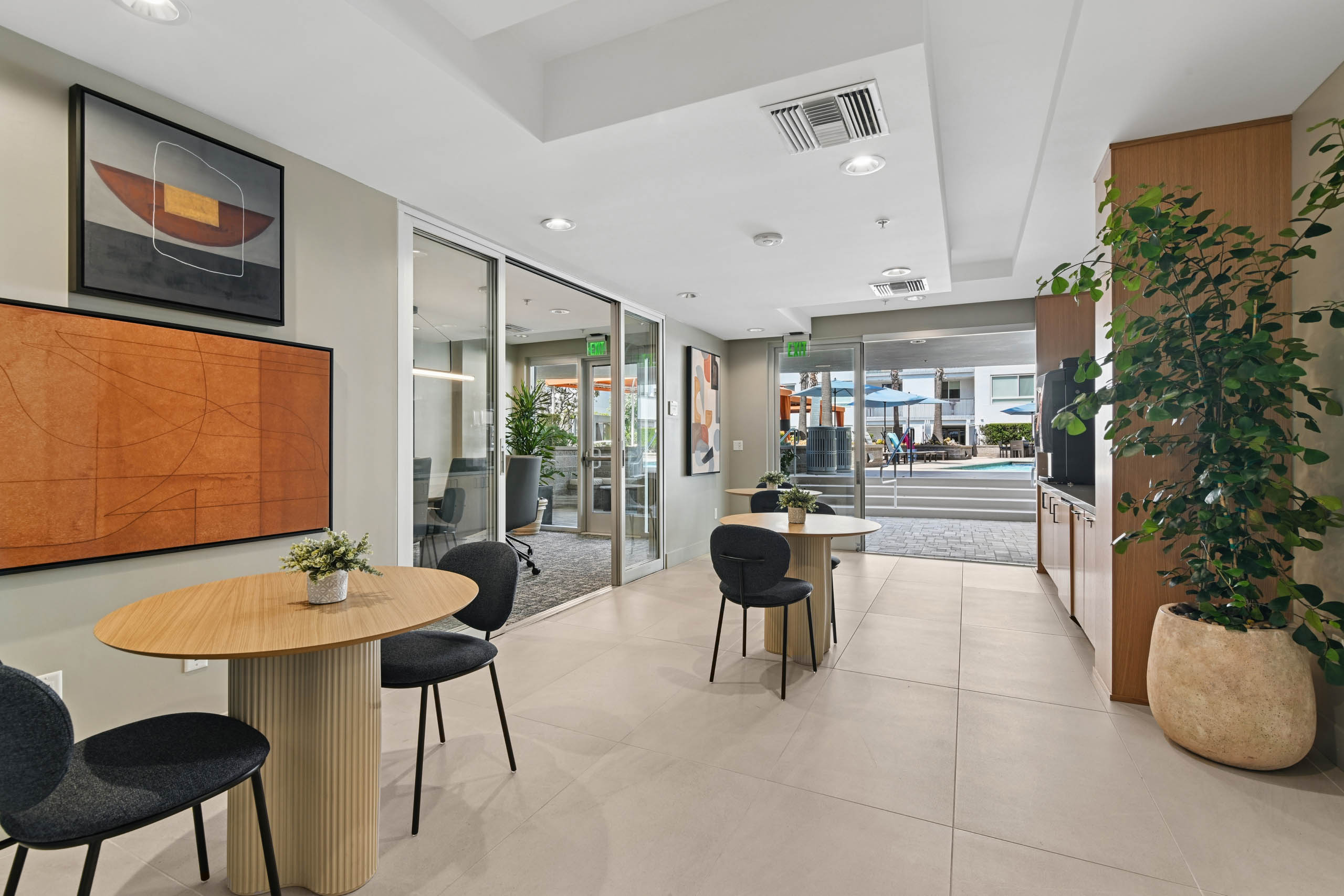Social Hub at AMLI Lex on Orange with round wood tables, dark upholstered chairs, abstract wall art, and wood cabinetry, next to a glass-walled conference room, with glass doors opening to the pool area.