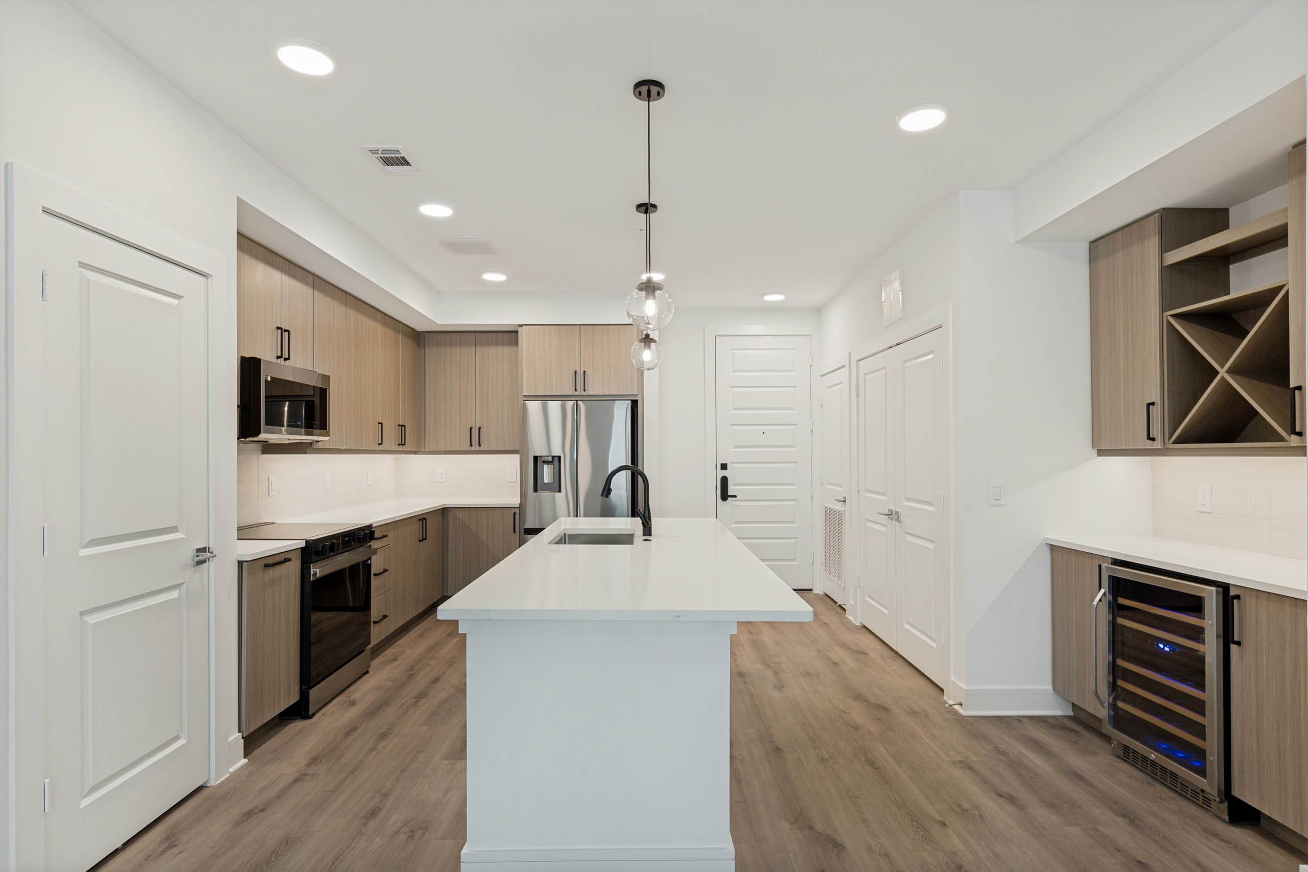 Spacious kitchen with large island, stainless steel appliances, wine refrigerator, built-in storage, pendant lighting, and wood flooring.