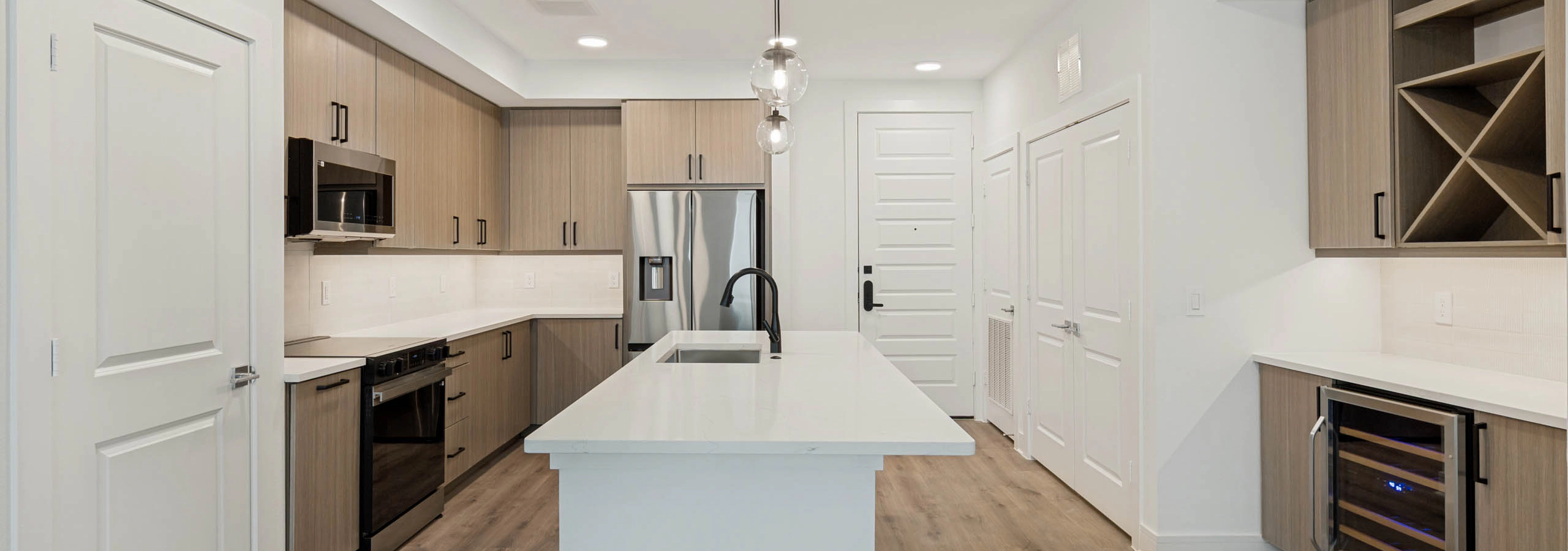 Spacious kitchen with large island, stainless steel appliances, wine refrigerator, built-in storage, pendant lighting, and wood flooring.