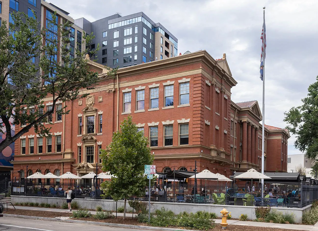 The brick covered exterior of Schoolyard Beer Garden housed in the historic Evans School with picnic tables and umbrellas outside on the patio
