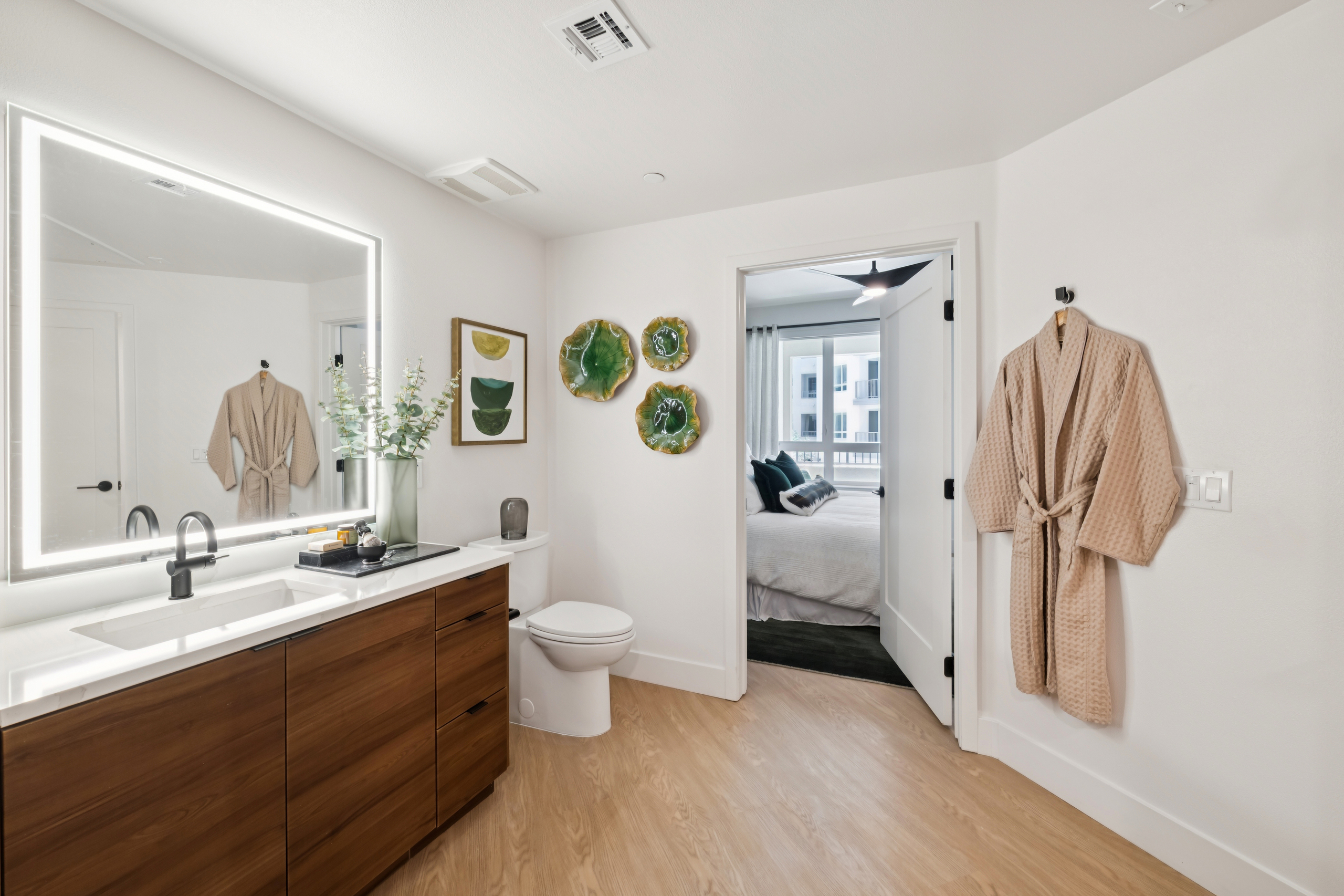 Modern bathroom with glass-enclosed shower, wood vanity, illuminated mirror, white countertop, black fixtures, towel bar, and walk-in closet.