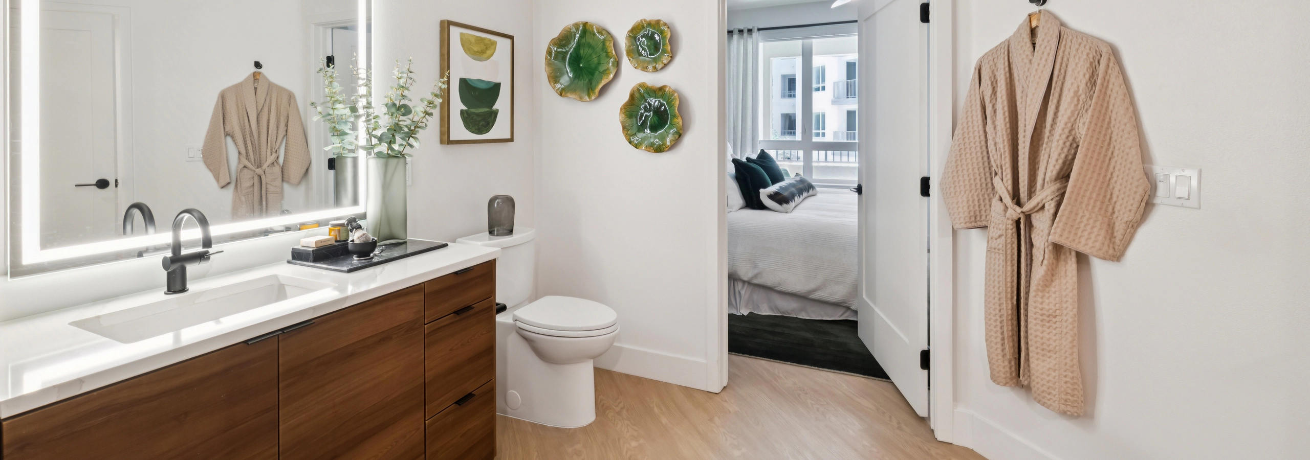 Modern bathroom with glass-enclosed shower, wood vanity, illuminated mirror, white countertop, black fixtures, towel bar, and walk-in closet.