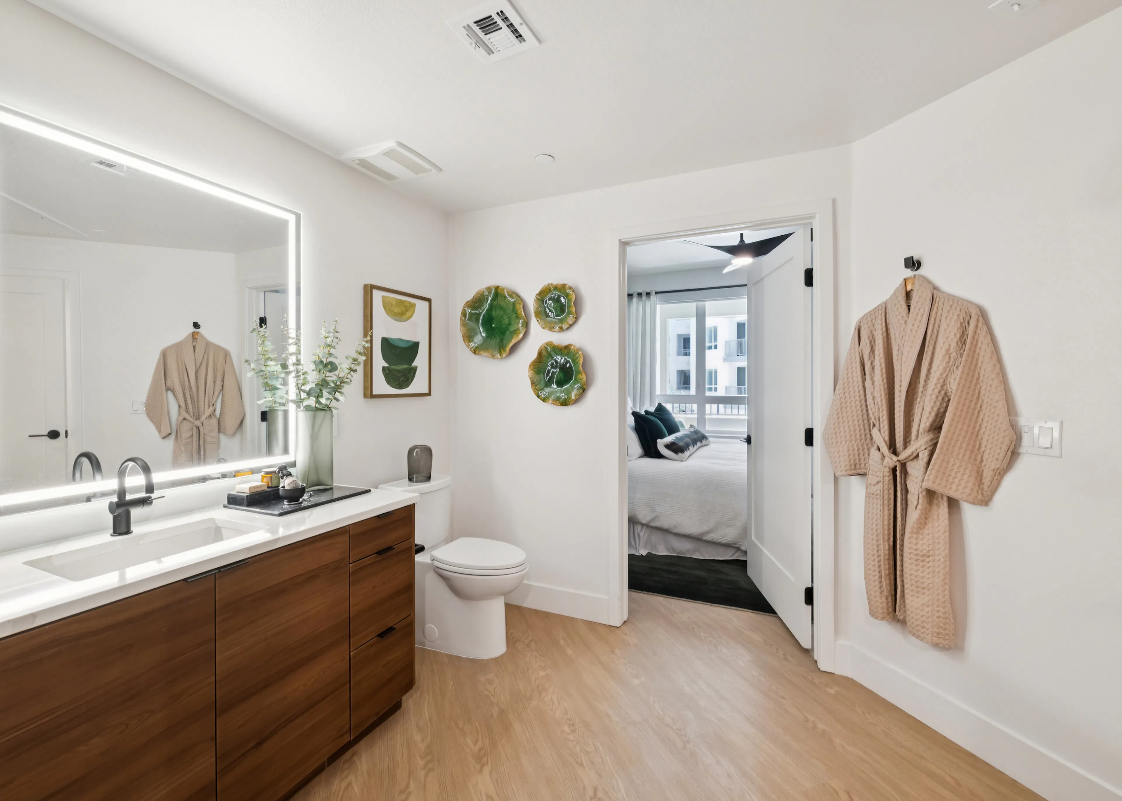 Modern bathroom with glass-enclosed shower, wood vanity, illuminated mirror, white countertop, black fixtures, towel bar, and walk-in closet.