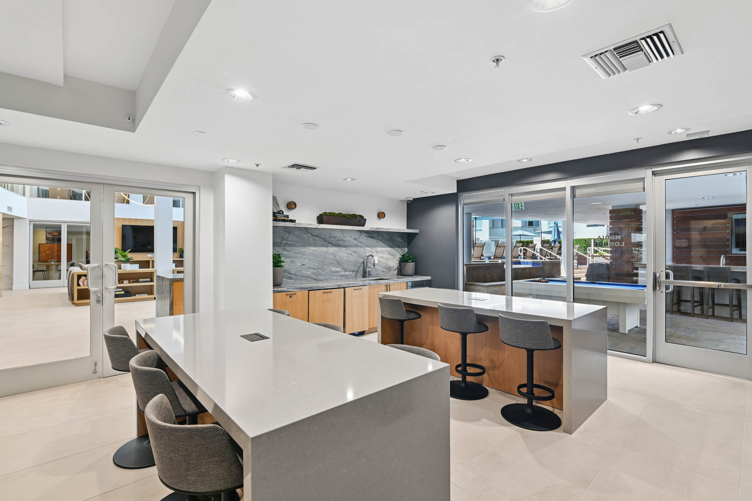 Media lounge at AMLI Lex on Orange with two counter-height tables with gray stools, a counter with sink, stone backsplash, and wood cabinets, and glass doors to an outdoor area with a pool table.
