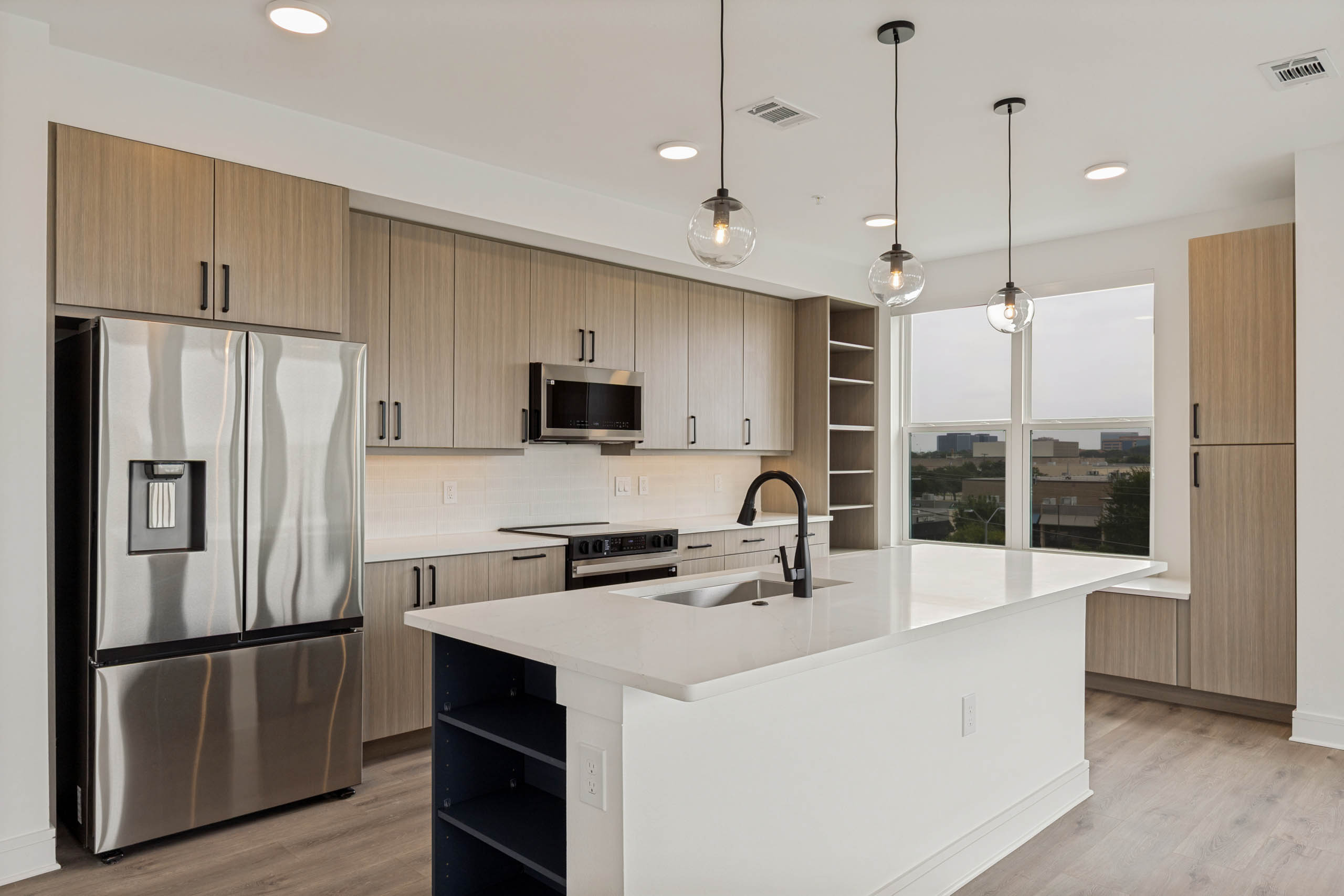 Modern kitchen with quartz island, stainless steel appliances, wood-style cabinetry, pendant lighting, open shelving, and large windows.
