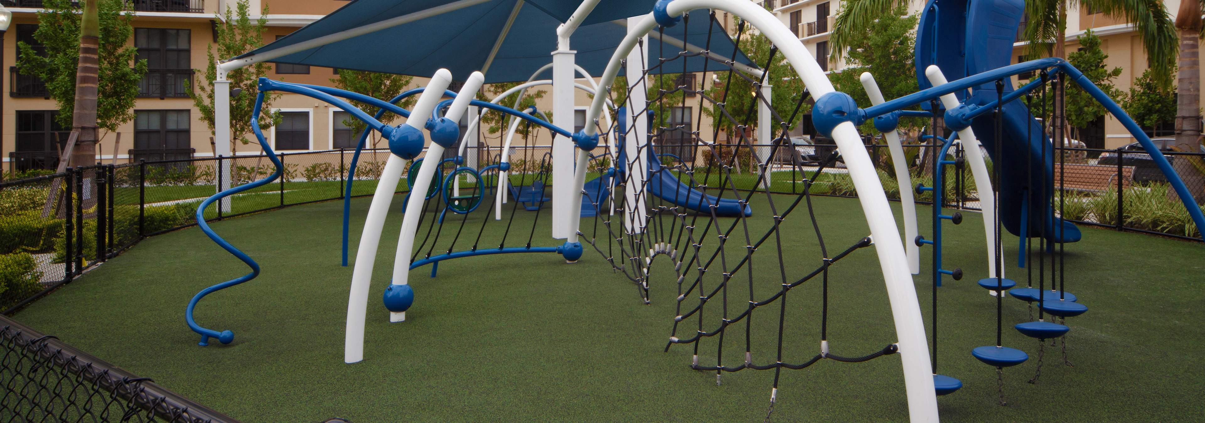Modern Children’s covered playground at AMLI Dadeland with blue and white recreational equipment on faux grass foundation