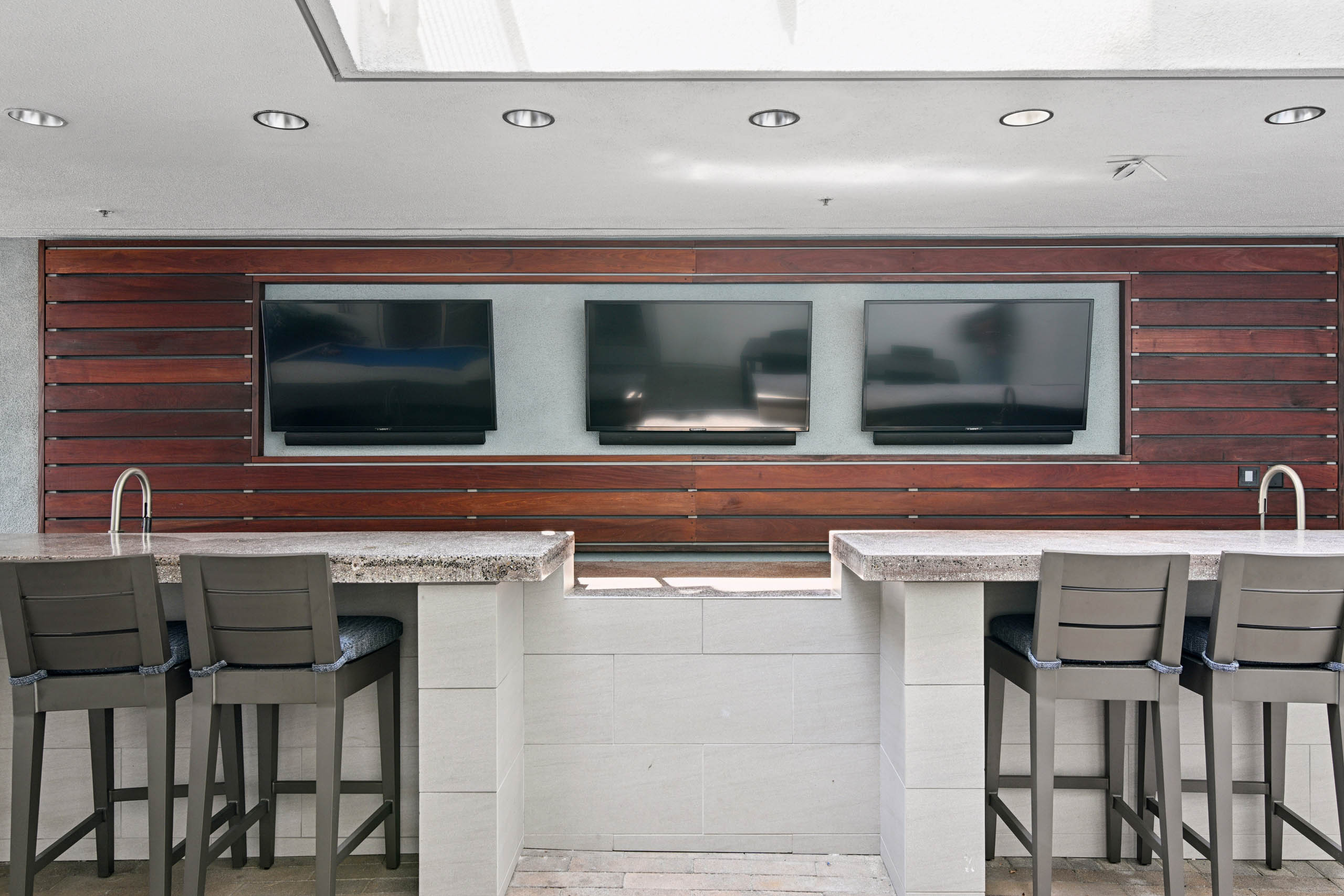 Covered outdoor bar at AMLI Lex on Orange with stone counters, two sinks, cushioned bar stools, and three wall-mounted TVs set into wood paneling.