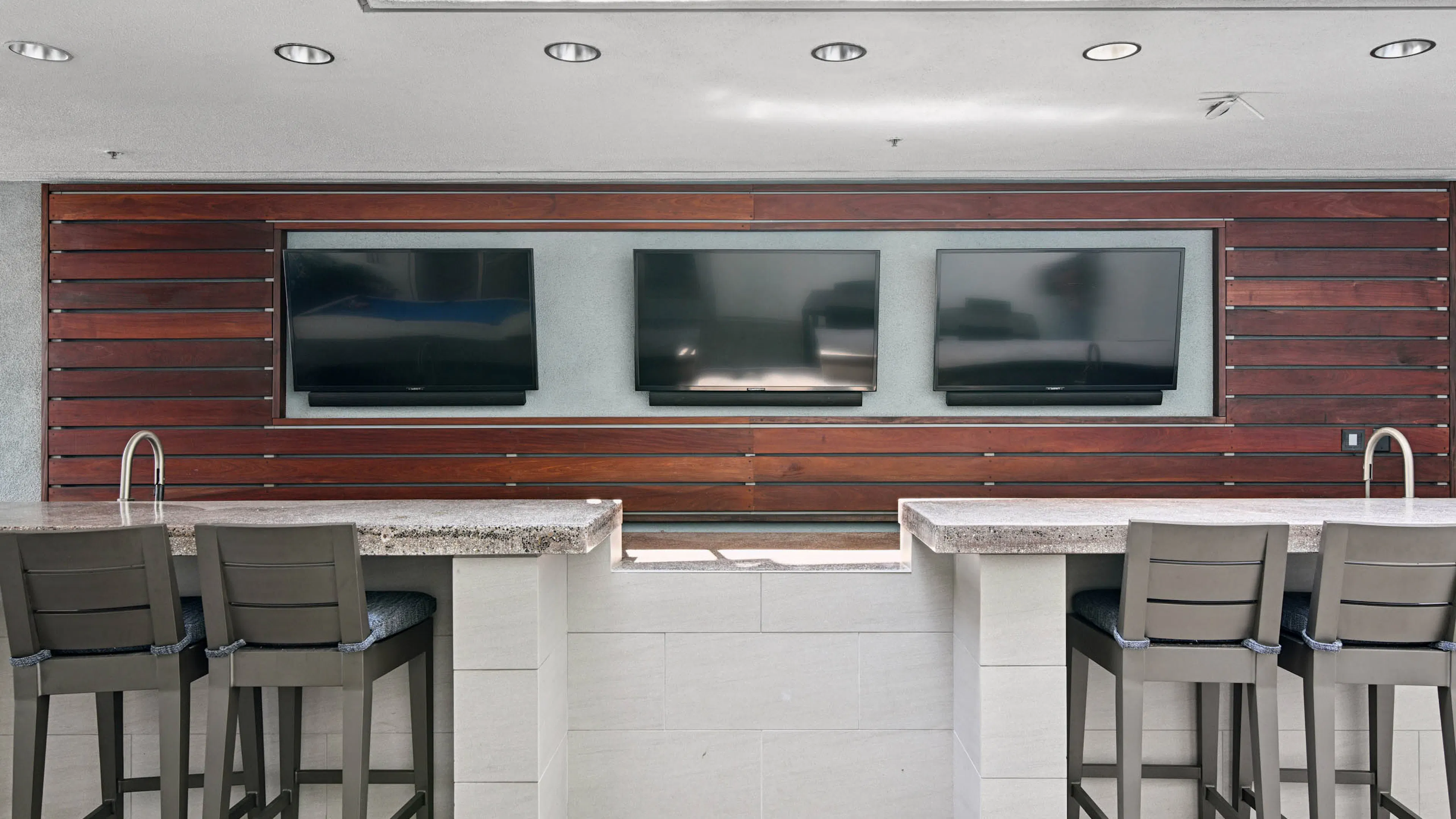 Covered outdoor bar at AMLI Lex on Orange with stone counters, two sinks, cushioned bar stools, and three wall-mounted TVs set into wood paneling.