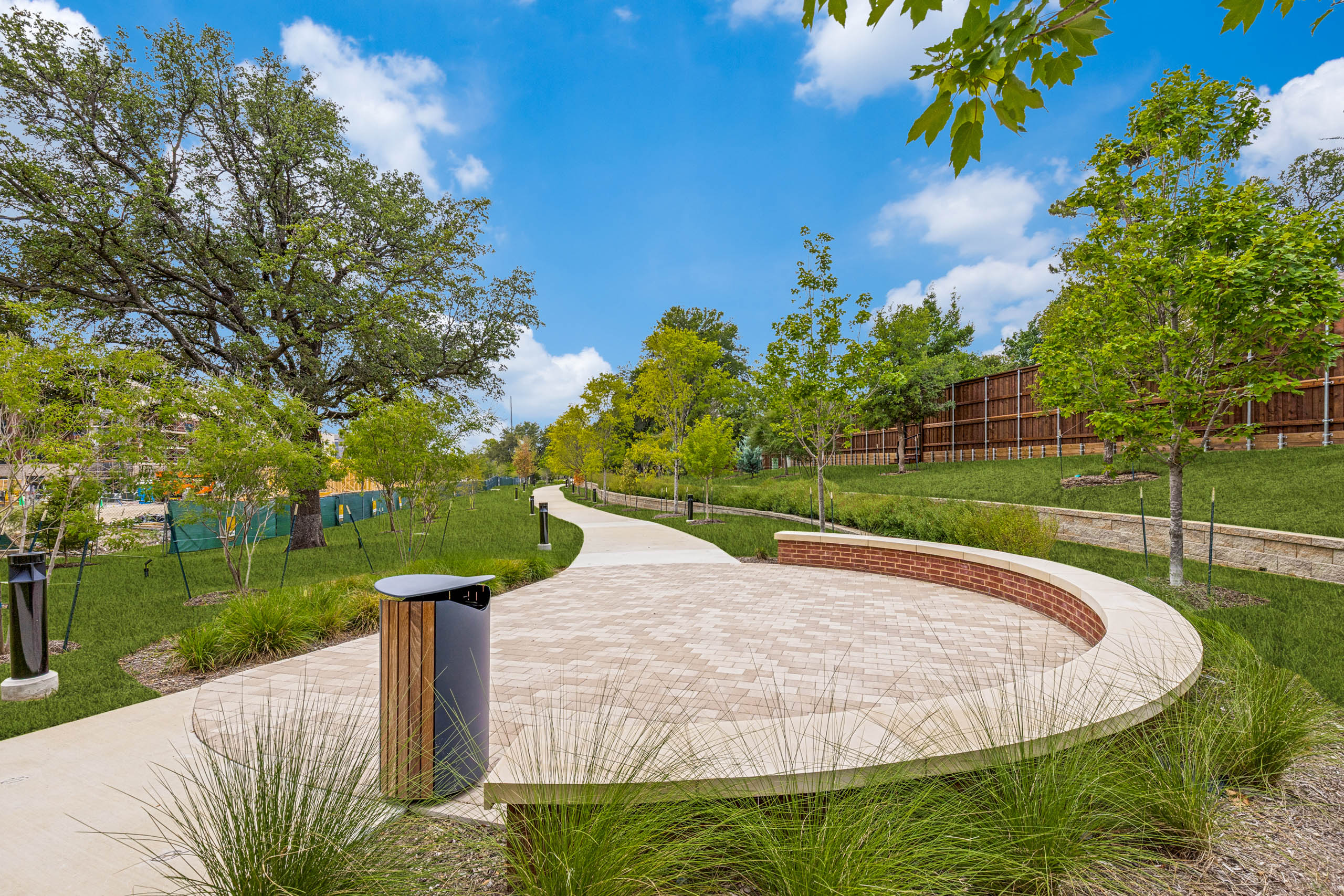 Outdoor gathering area with a curved seating wall, paved plaza, landscaped grounds, mature greenery, and a winding walking trail at AMLI Treehouse.