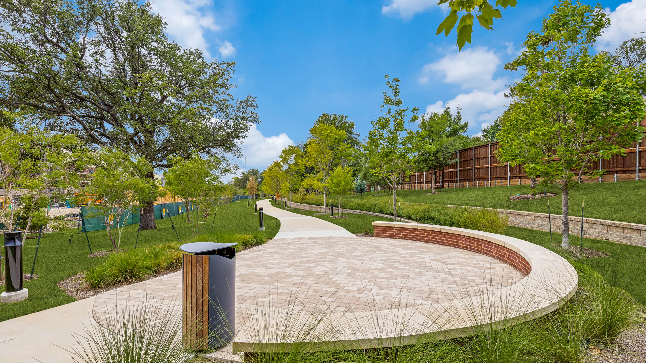 Outdoor gathering area with a curved seating wall, paved plaza, landscaped grounds, mature greenery, and a winding walking trail at AMLI Treehouse.