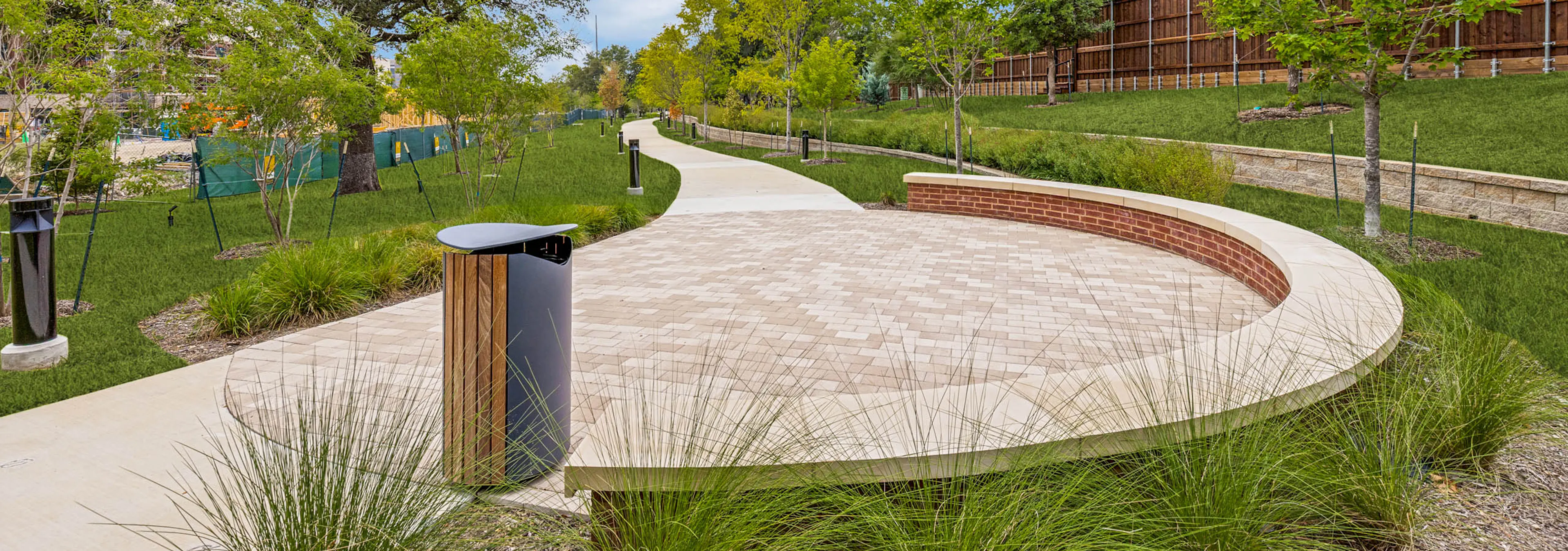 Outdoor gathering area with a curved seating wall, paved plaza, landscaped grounds, mature greenery, and a winding walking trail at AMLI Treehouse.