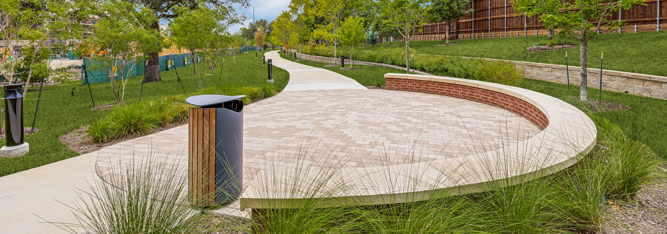 Outdoor gathering area with a curved seating wall, paved plaza, landscaped grounds, mature greenery, and a winding walking trail at AMLI Treehouse.