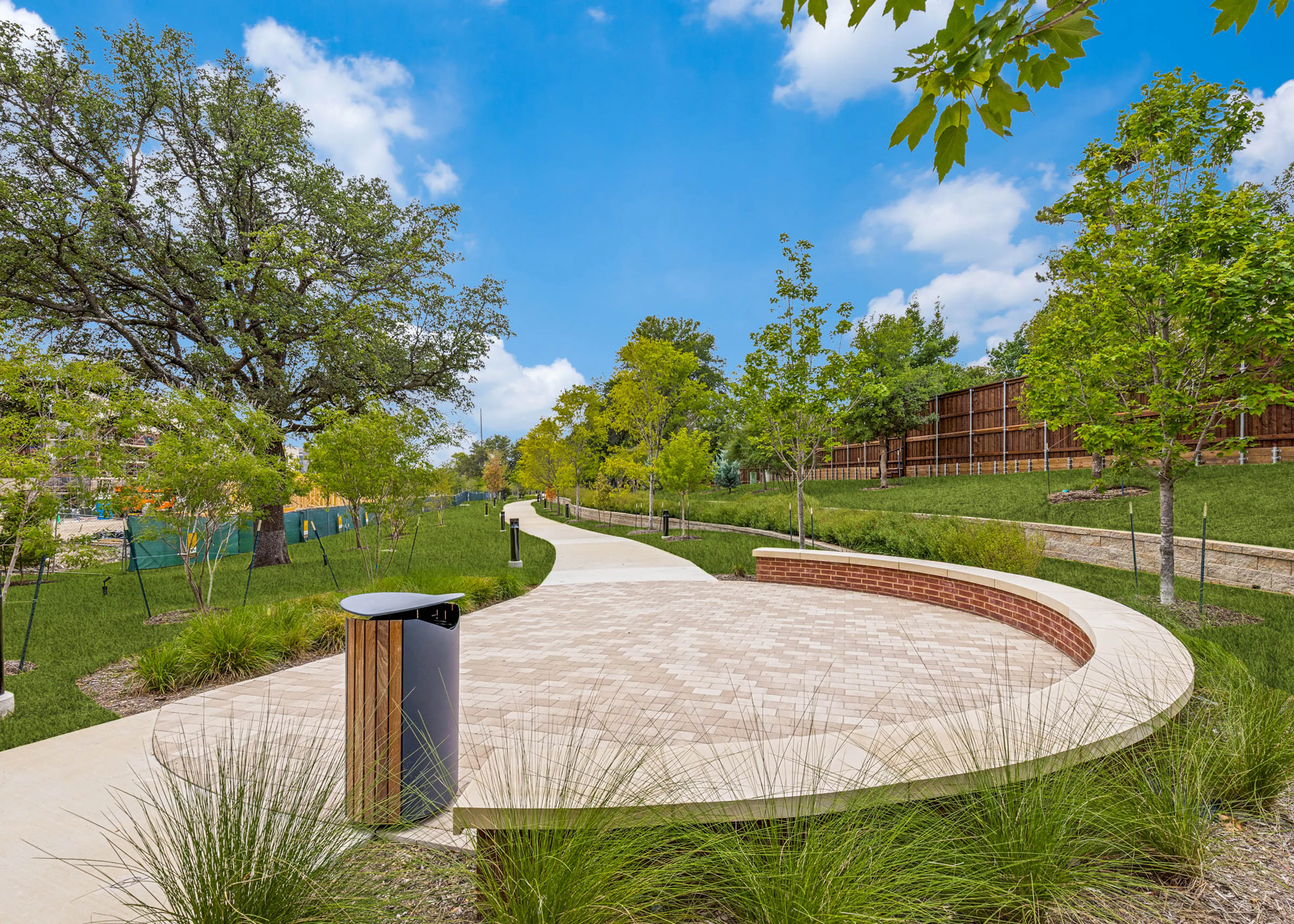Outdoor gathering area with a curved seating wall, paved plaza, landscaped grounds, mature greenery, and a winding walking trail at AMLI Treehouse.