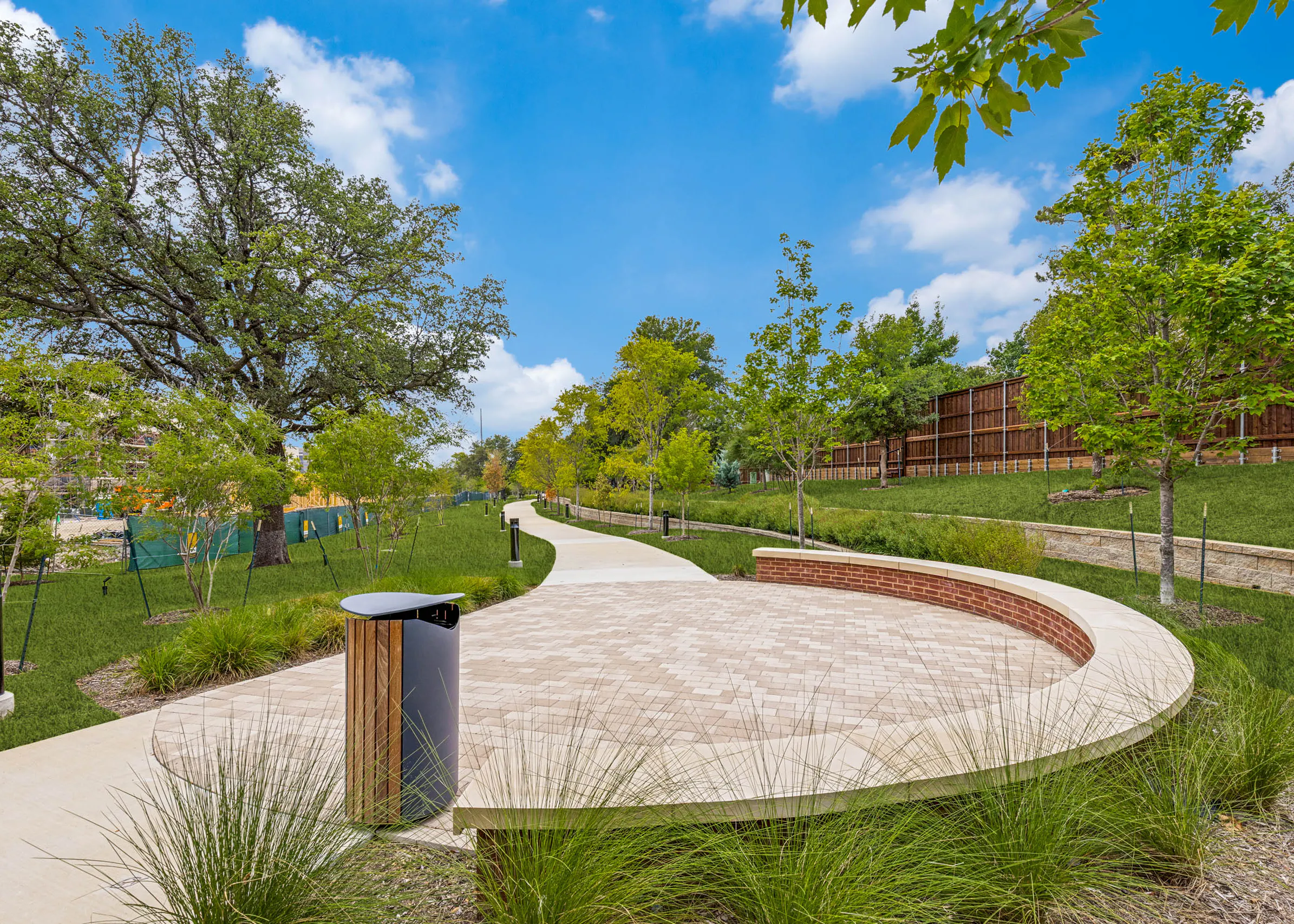 Outdoor gathering area with a curved seating wall, paved plaza, landscaped grounds, mature greenery, and a winding walking trail at AMLI Treehouse.