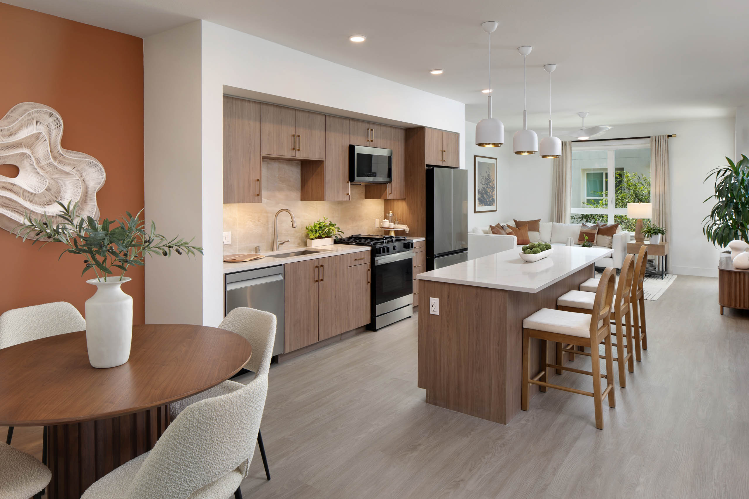 Open-concept kitchen and living area with wood cabinets, white island, bar stools, dining table, pendant lights, plants, and large window.