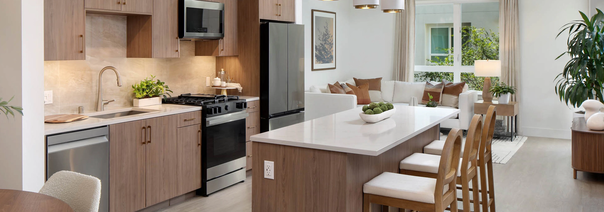 Open-concept kitchen and living area with wood cabinets, white island, bar stools, dining table, pendant lights, plants, and large window.