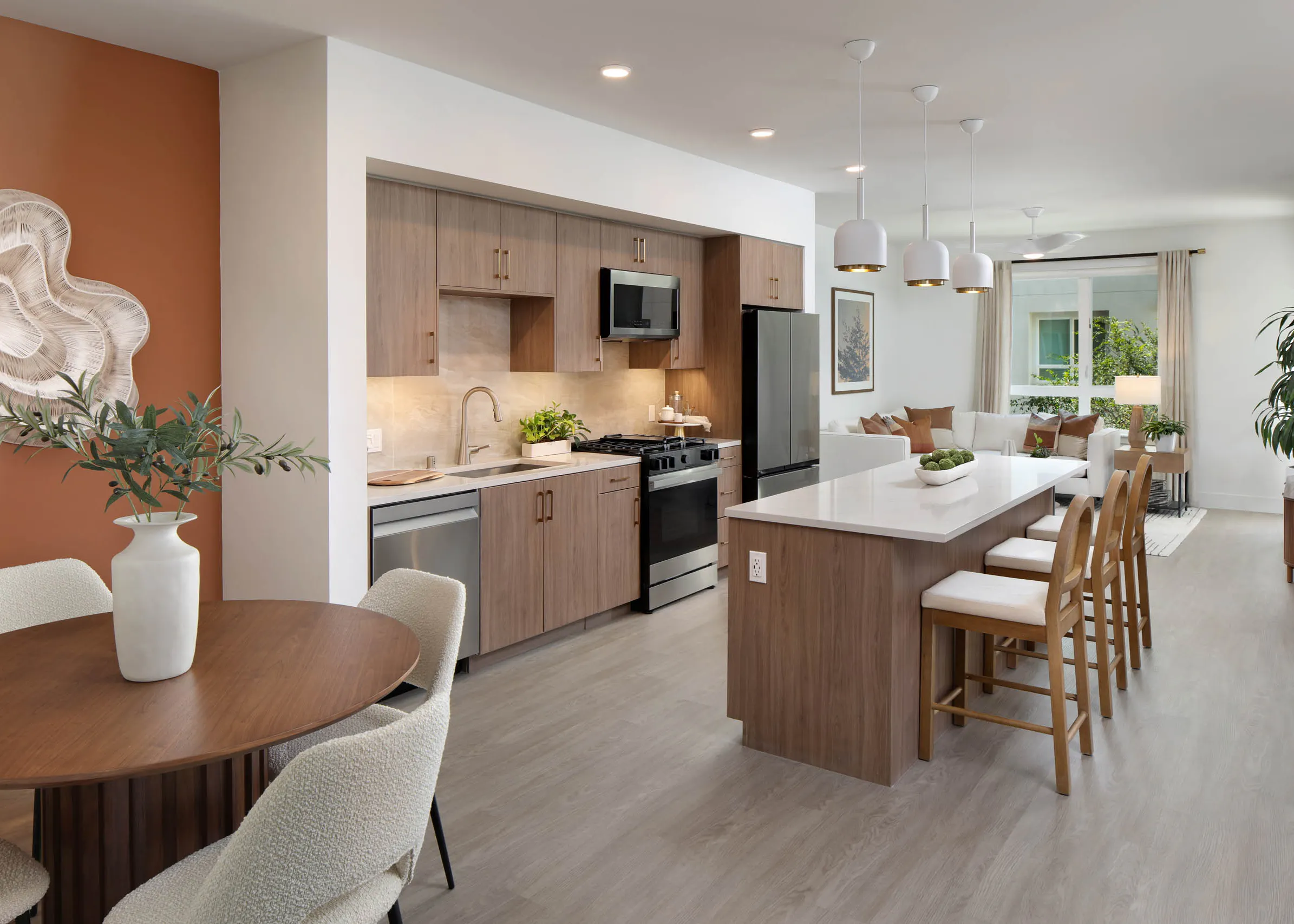 Open-concept kitchen and living area with wood cabinets, white island, bar stools, dining table, pendant lights, plants, and large window.