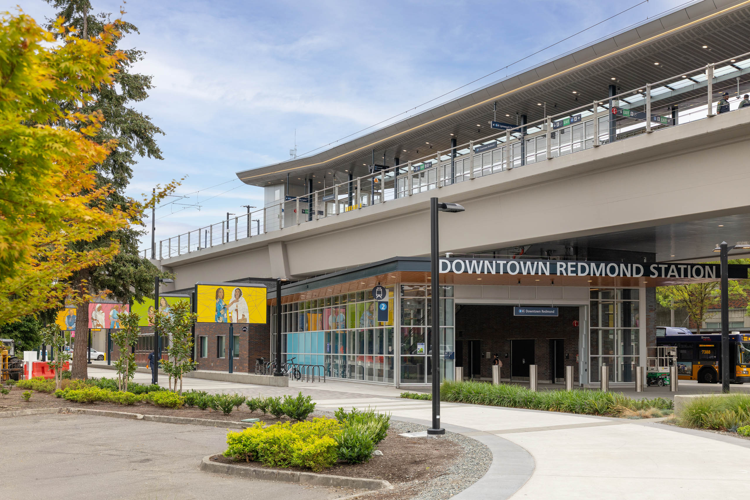 Downtown Redmond Station entrance with colorful mural artwork, landscaping, and the elevated light rail platform above, located near AMLI Redmond Way.